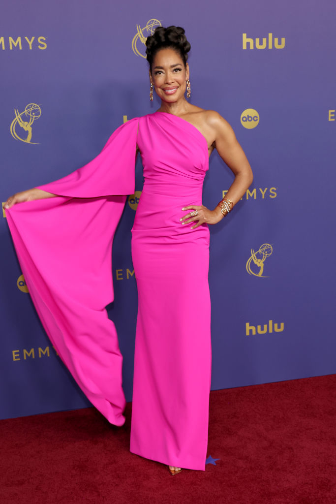 A woman stands on the red carpet at the Emmys, showcasing a one-shoulder, floor-length gown with a flowing sleeve. She is smiling and posing for the camera