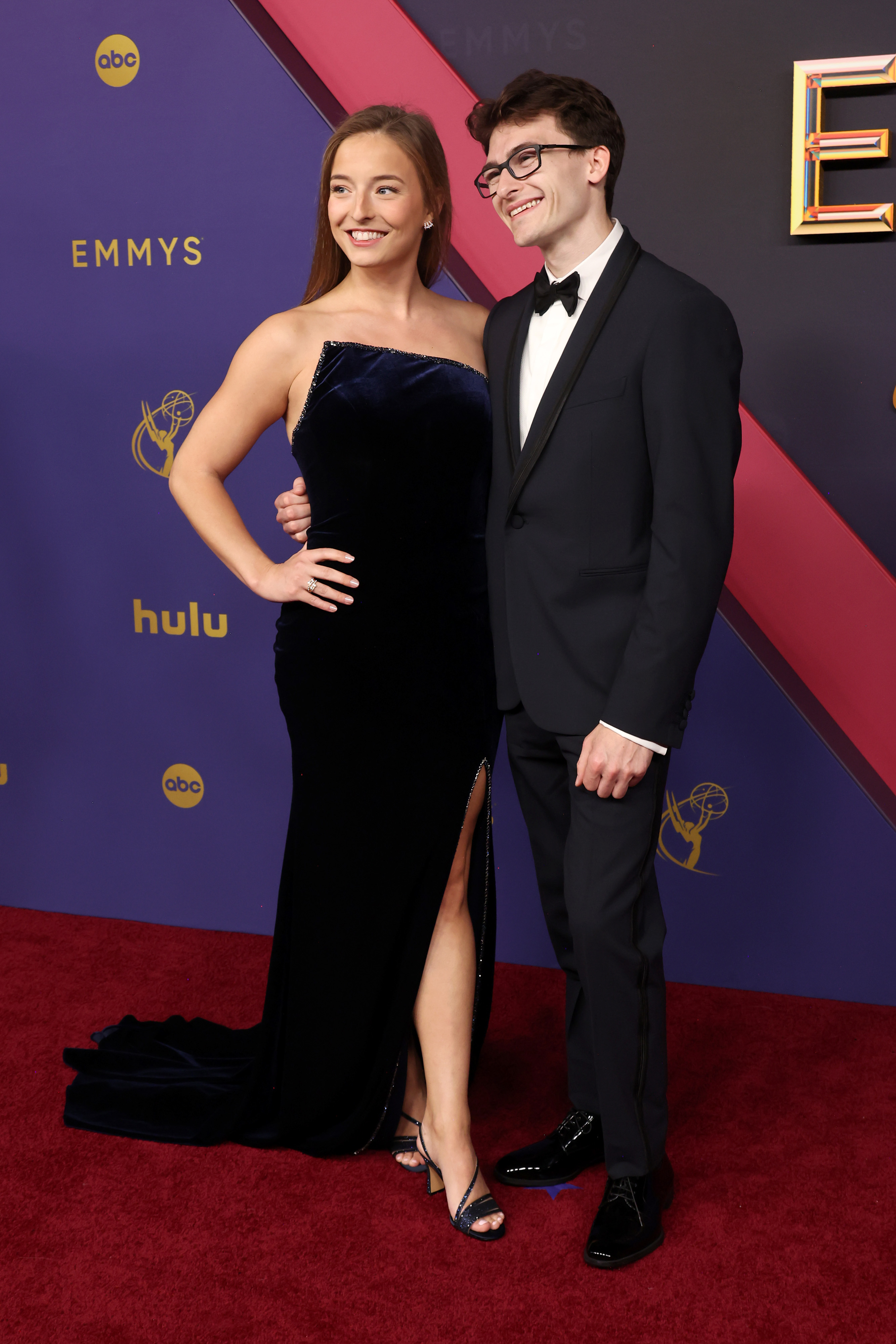 Stephen Nedoroscik in a black tuxedo and bow tie with glasses and Tess McCracken in a black strapless gown with a thigh-high slit smile together at the Emmys red carpet