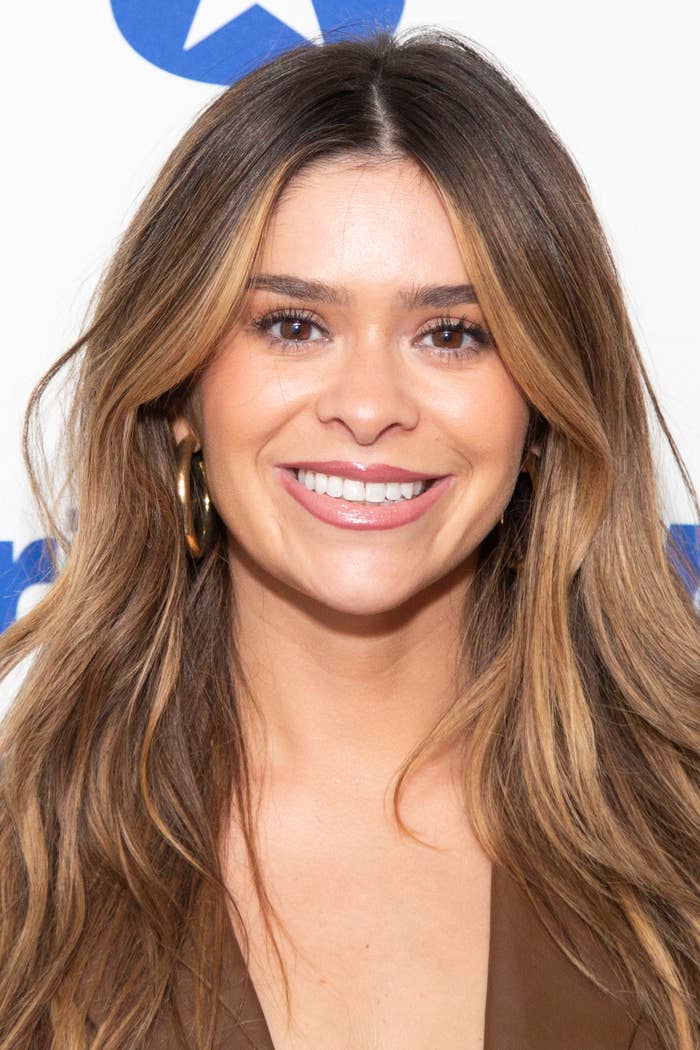 A woman with long, wavy hair smiles warmly at the camera, wearing large hoop earrings and a stylish outfit