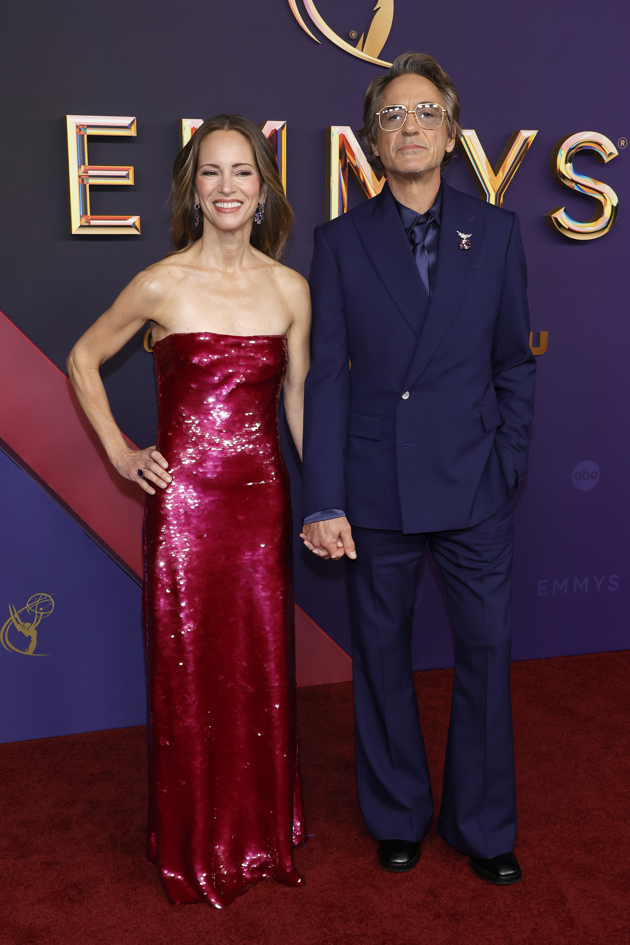 Susan Downey and Robert Downey Jr. pose on the red carpet at the Emmys. Susan wears a sparkling strapless dress, and Robert is in a sleek navy suit with sunglasses