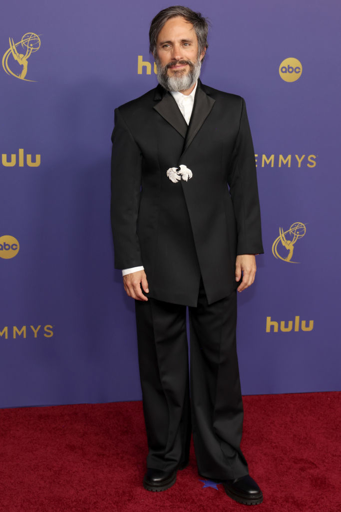 A man stands on a red carpet at an event, wearing a black suit with a white shirt and a unique decorative brooch