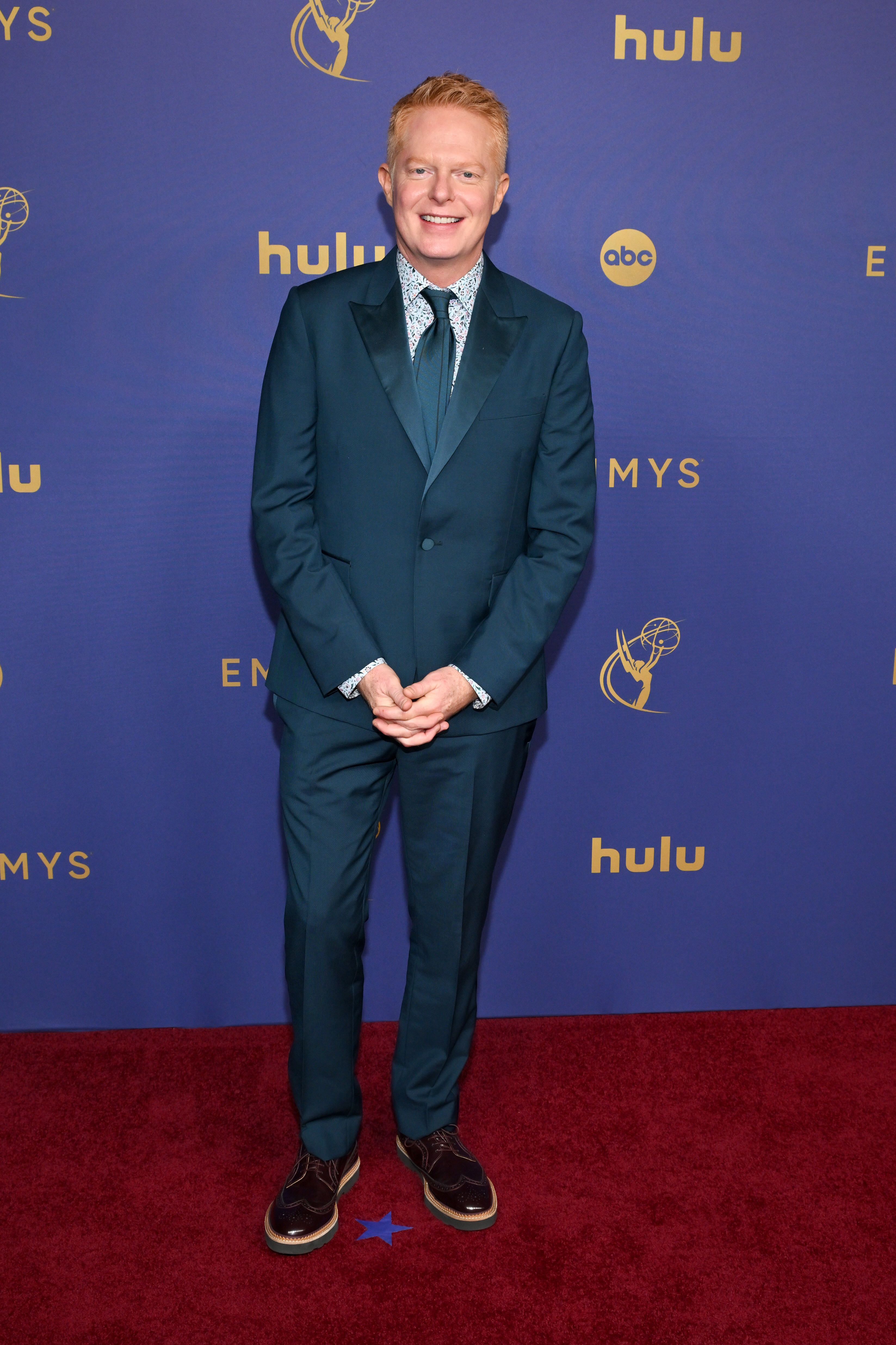Jesse Tyler Ferguson smiles while wearing a suit with tie and shoes on the red carpet at the Emmy Awards