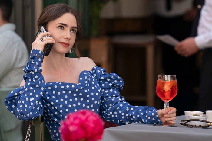 Lily Collins in an off-shoulder, polka dot dress, holding a phone to her ear and a drink in her other hand, seated at an outdoor café