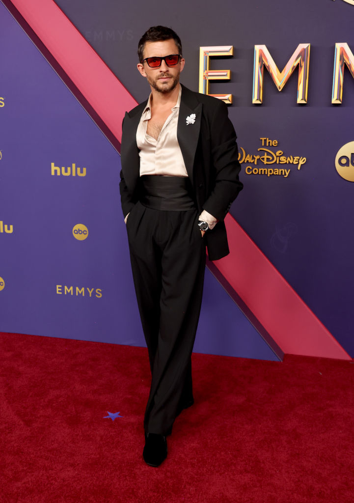 Hombre en la alfombra roja de los Emmys con gafas de sol, camisa abierta, y esmoquin sin corbata posando con las manos en los bolsillos