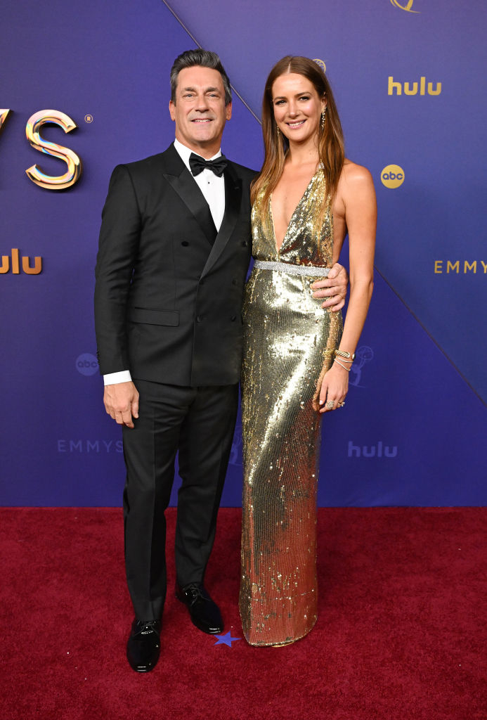 Two celebrities in formal attire pose on the red carpet at the Emmys. The man wears a black tuxedo, and the woman wears a gold, sleeveless gown