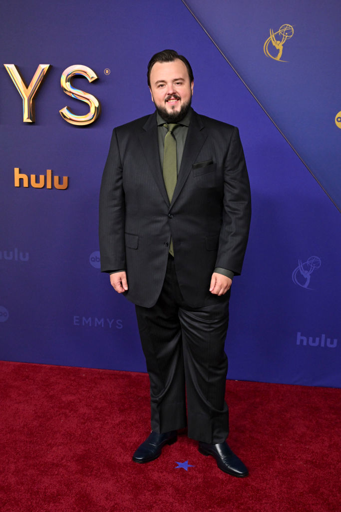 John Bradley in a tailored black suit, green shirt, and black tie, posing on the red carpet at the Emmys event