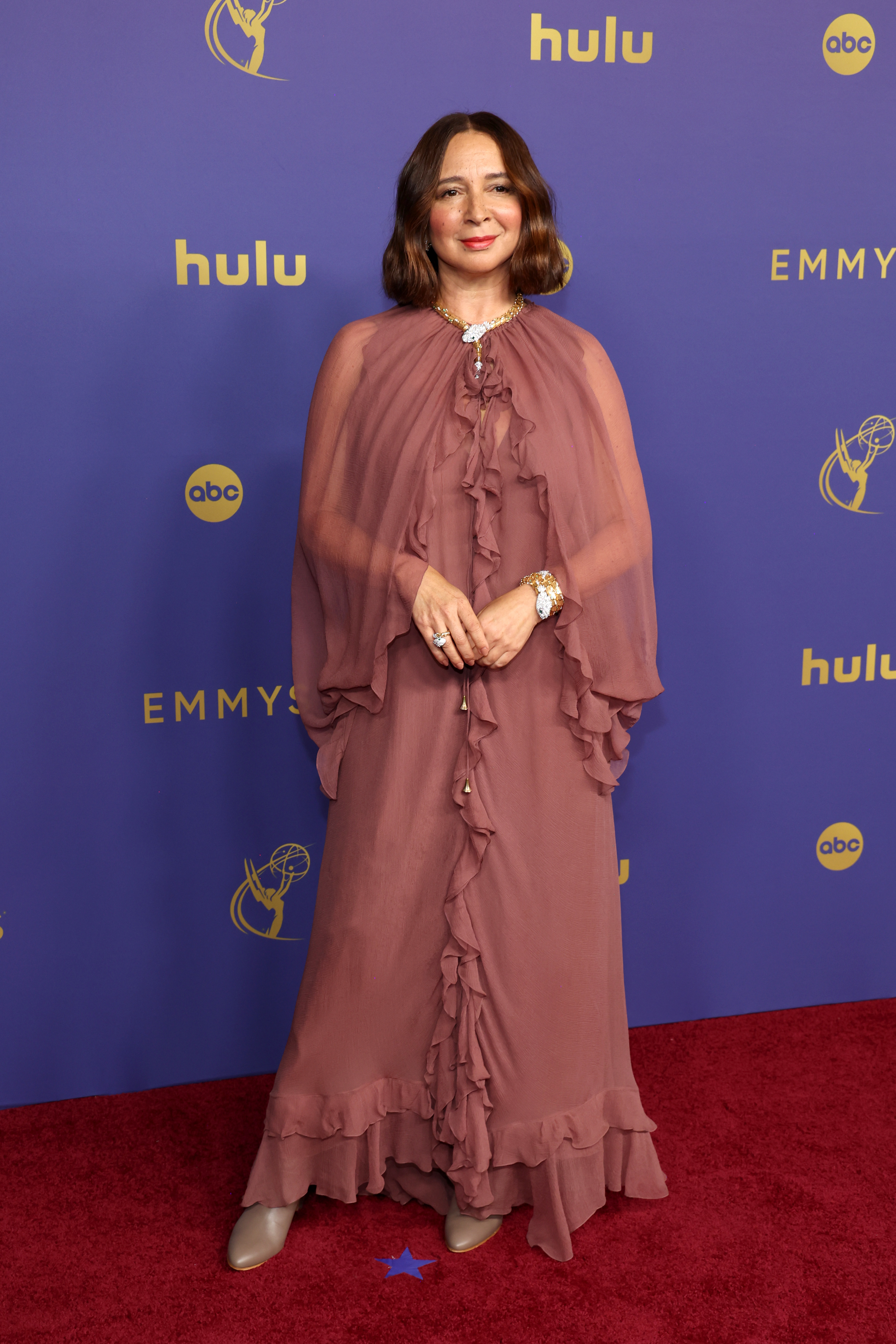 Maya Rudolph on the red carpet at the Emmys, wearing a flowing, ruffled dress with long, sheer sleeves