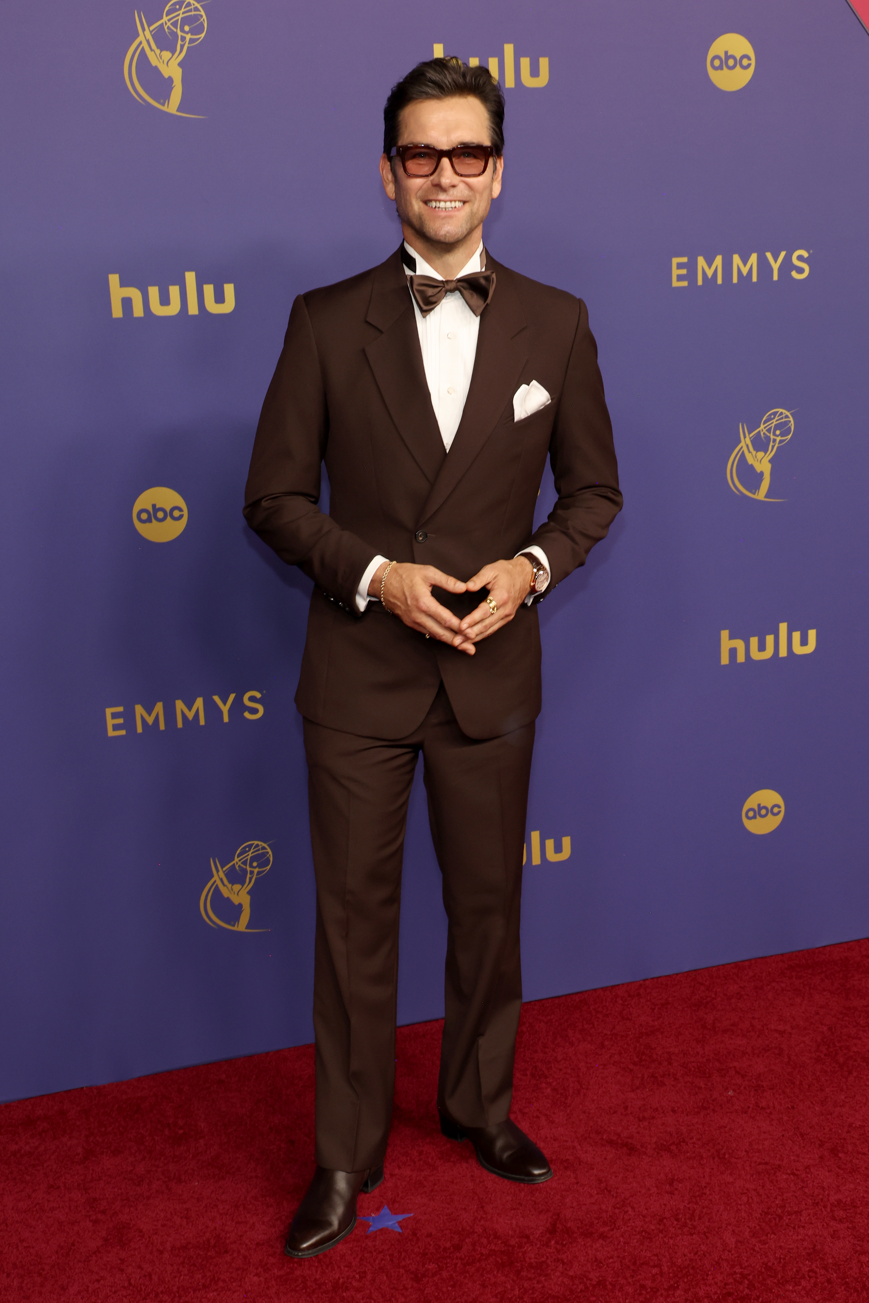 Antony Starr posing on the red carpet at the Emmys in a suit with a bow tie and glasses