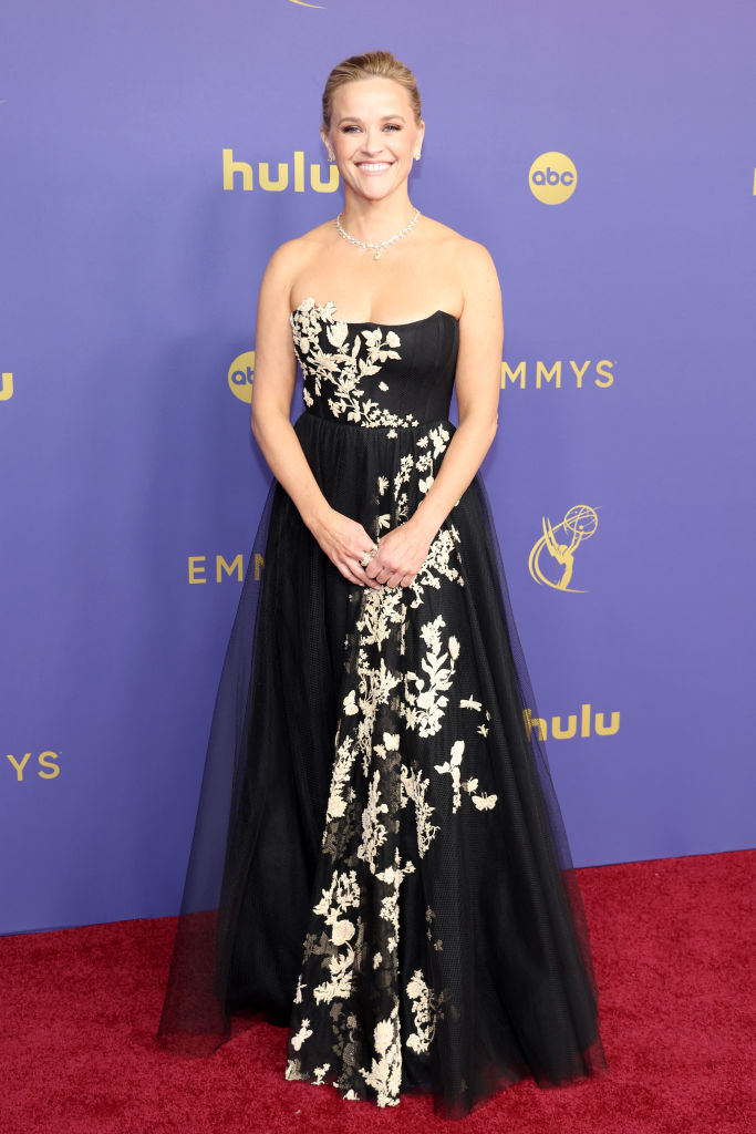 Reese Witherspoon on the red carpet, wearing a strapless black gown with white floral embroidery, smiling for the cameras at the Emmys