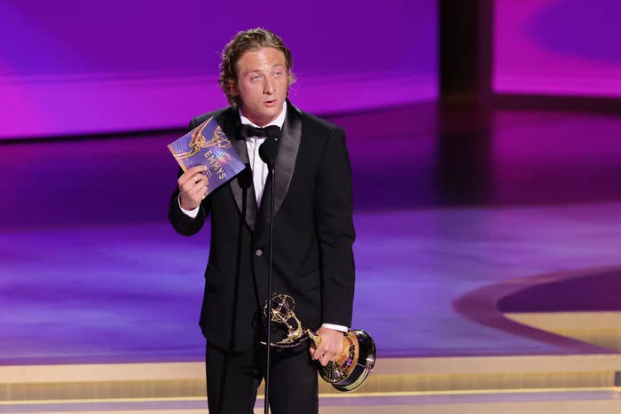 Jeremy Allen White gives a speech on stage while holding an Emmy Award in one hand and a winner's envelope in the other. He is dressed in a black suit