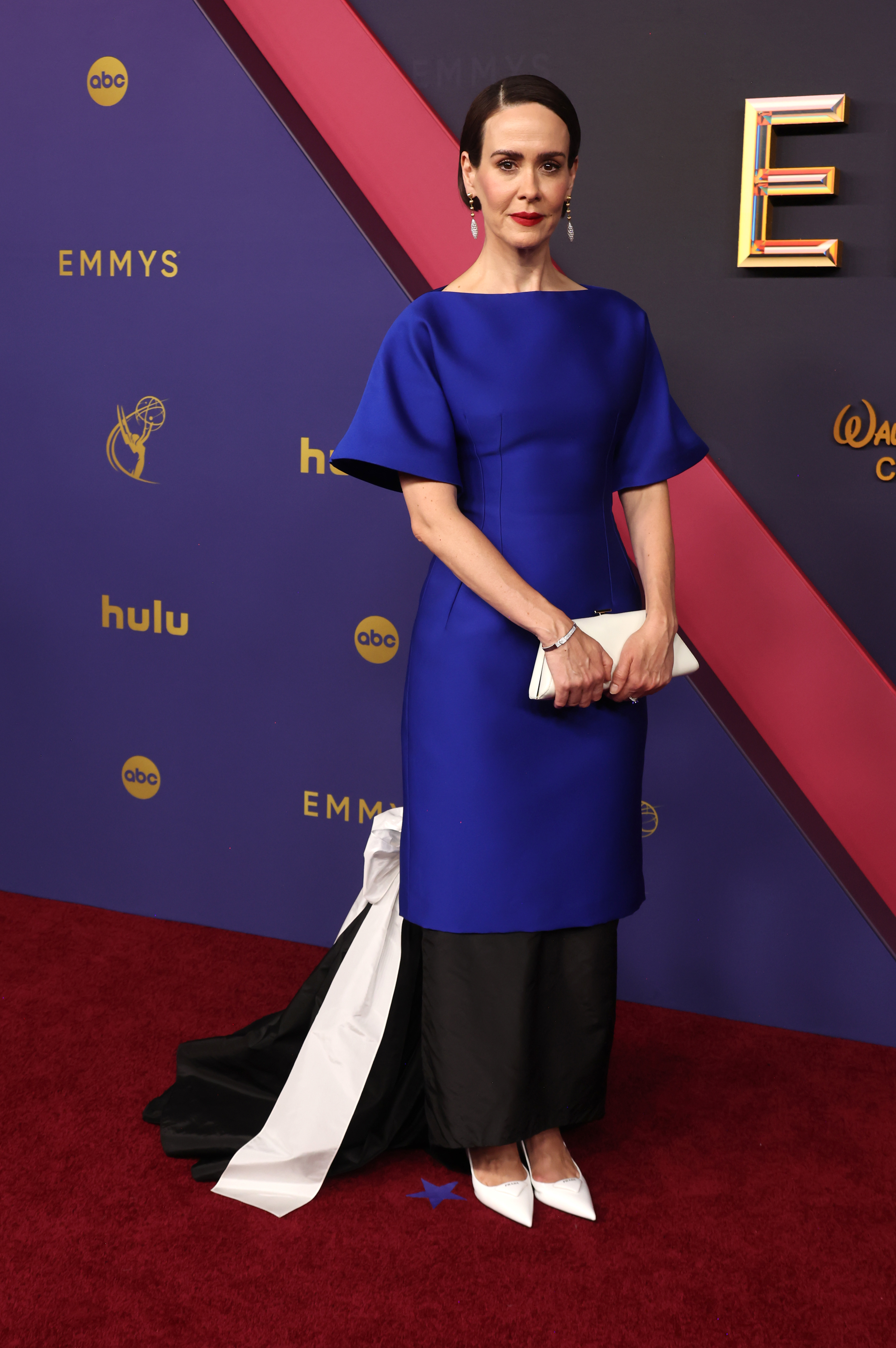Sarah Paulson on the red carpet at the Emmys, wearing a blue dress with statement sleeves and a black and white train, holding a white clutch