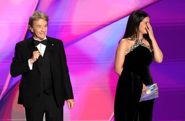 Martin Short in a tuxedo and Selena Gomez in an evening gown on stage at an event, with Selena covering her face while holding a card