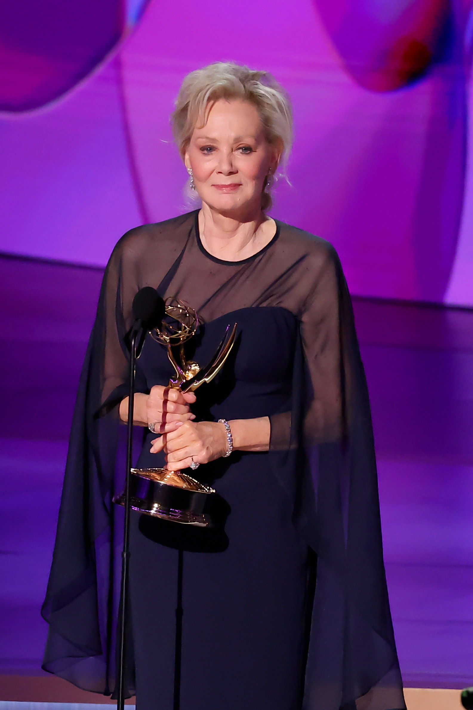 Jean Smart holding an Emmy Award on stage, wearing an elegant black gown with sheer sleeves