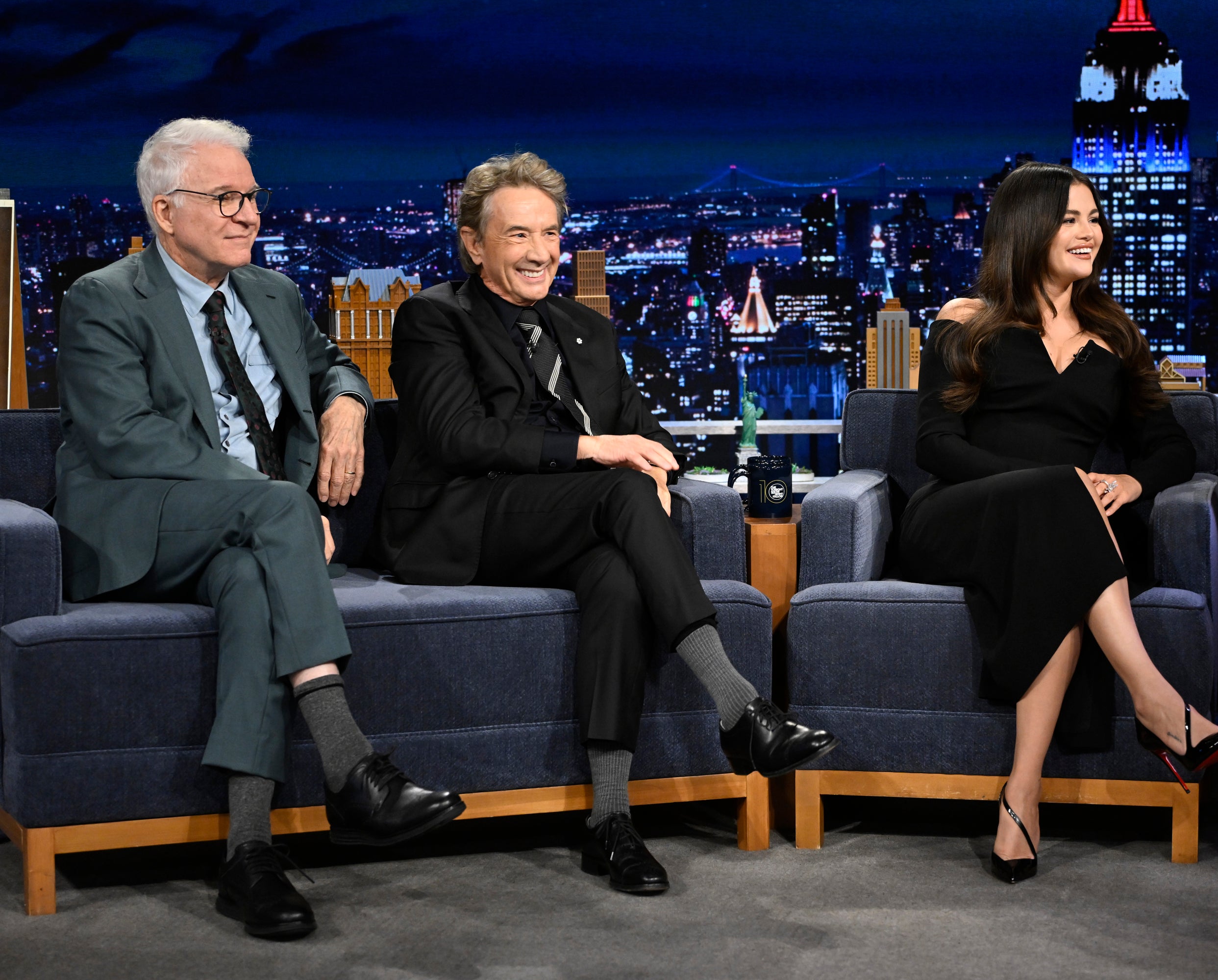 Steve Martin, Martin Short, and Selena Gomez are sitting on a talk show set, engaging in conversation, with a cityscape backdrop visible in the background