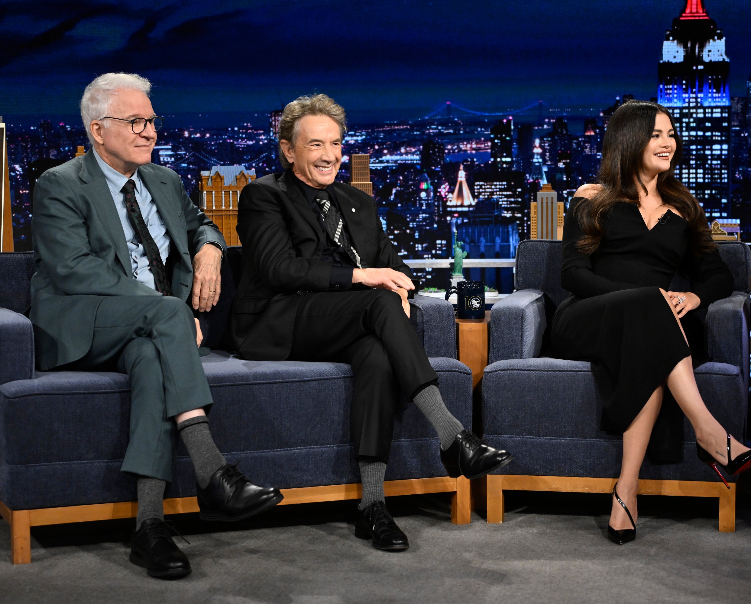 Steve Martin, Martin Short, and Selena Gomez are sitting on a talk show set, engaging in conversation, with a cityscape backdrop visible in the background
