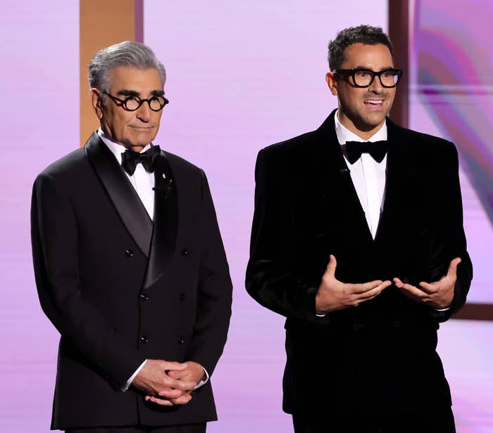 Eugene Levy and Dan Levy are standing side by side on stage in tuxedos, speaking and smiling during an awards ceremony