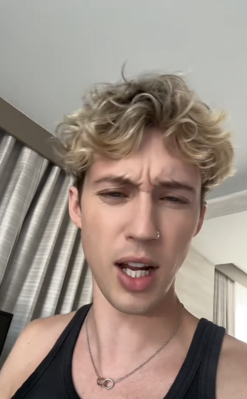 Troye Sivan gazes at the camera, wearing a black tank top and a silver necklace, with short curly hair