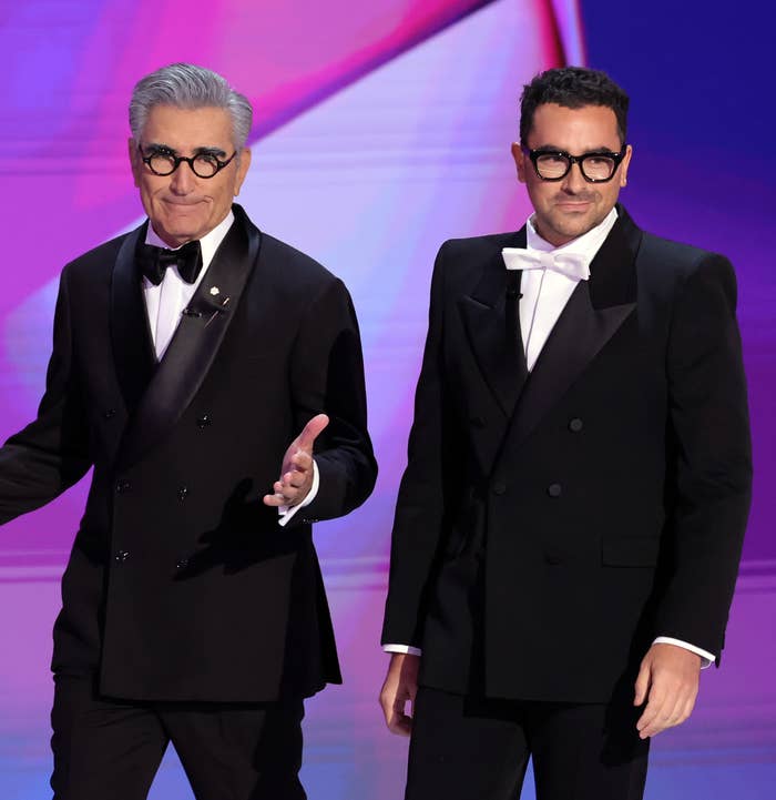 Eugene Levy and Dan Levy on stage in black tuxedos with bow ties. Eugene gestures with his hands while Dan stands with hands at his sides