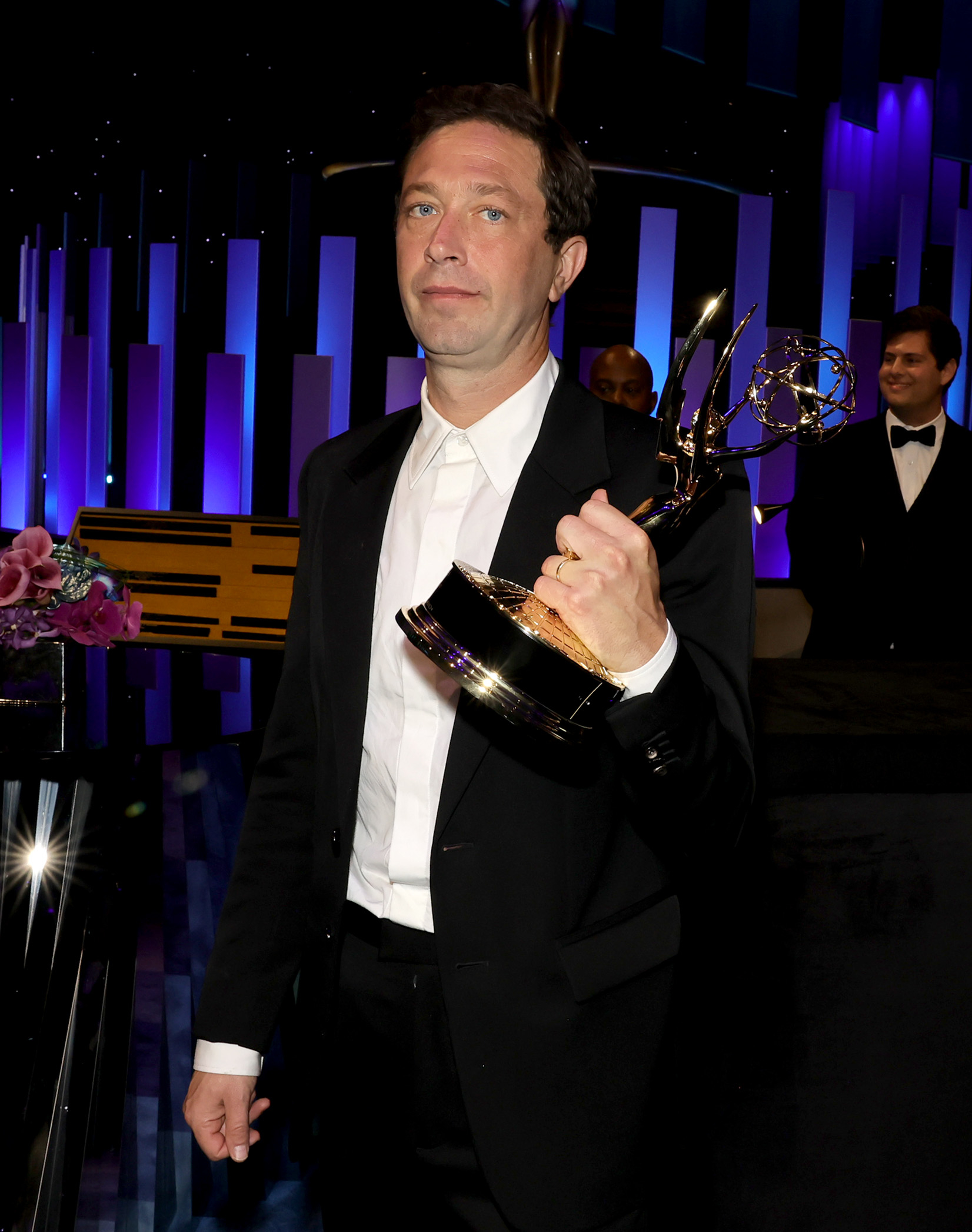 Ebon Moss-Bachrach in a dark suit holds an Emmy award on stage with others in the background during an award ceremony