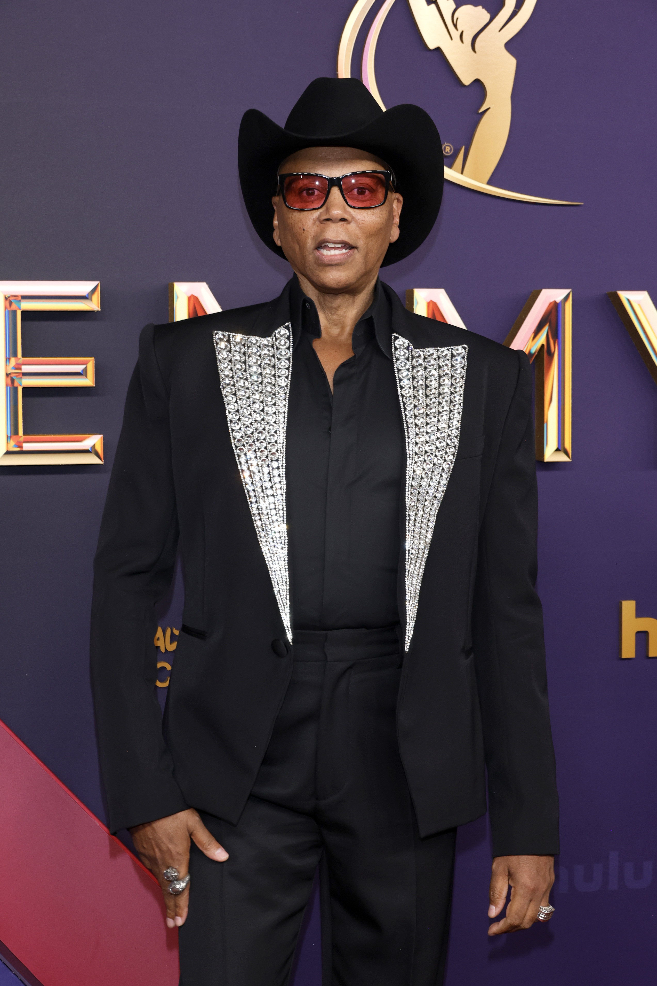 RuPaul in a dark cowboy hat, suit with sparkling silver lapels, and tinted sunglasses at an event