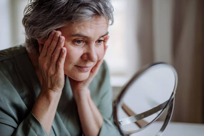 Older woman with short gray hair looking at her reflection in a mirror, gently holding her face with her hands