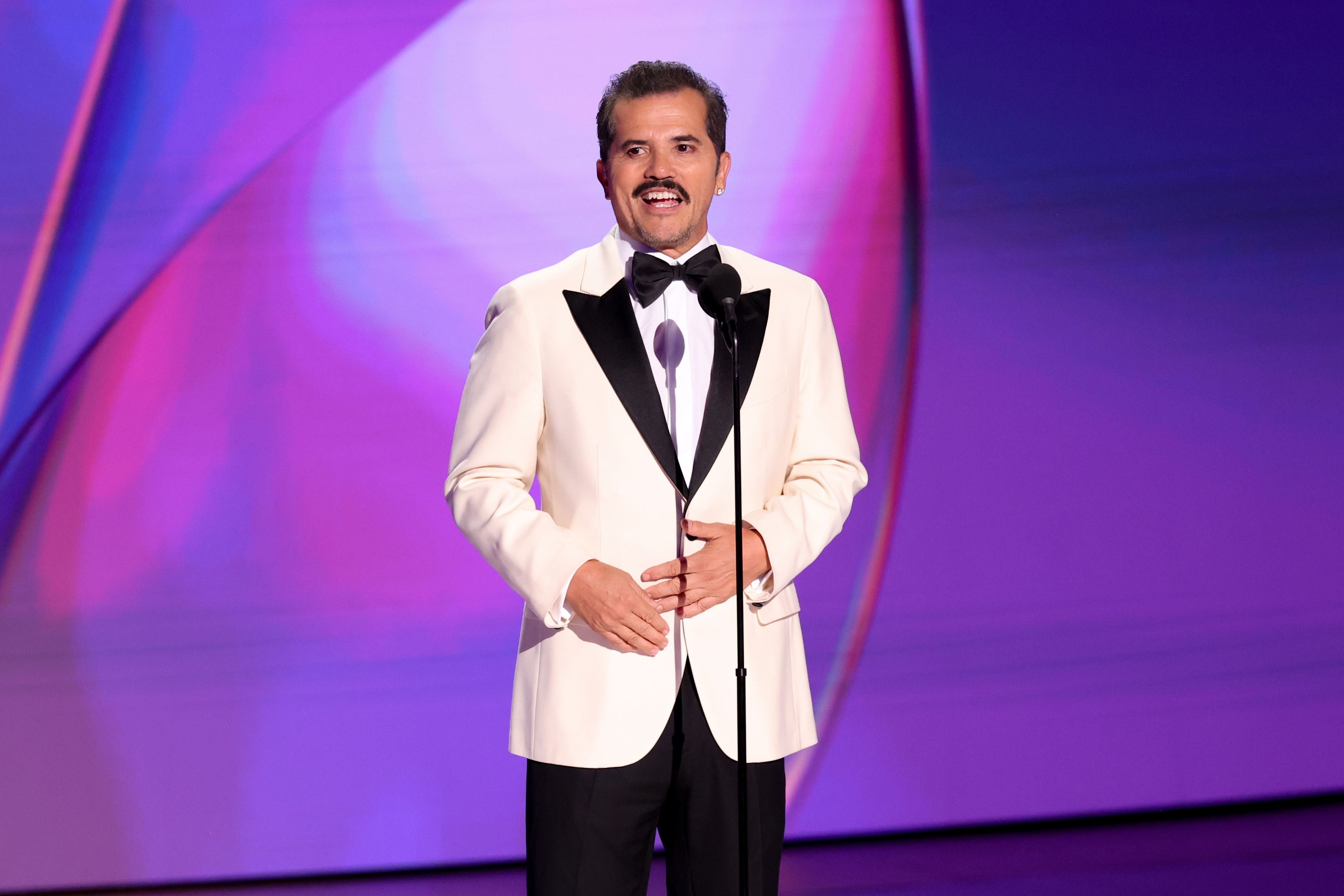 John Leguizamo stands on stage in a formal white tuxedo with black lapels, speaking into a microphone