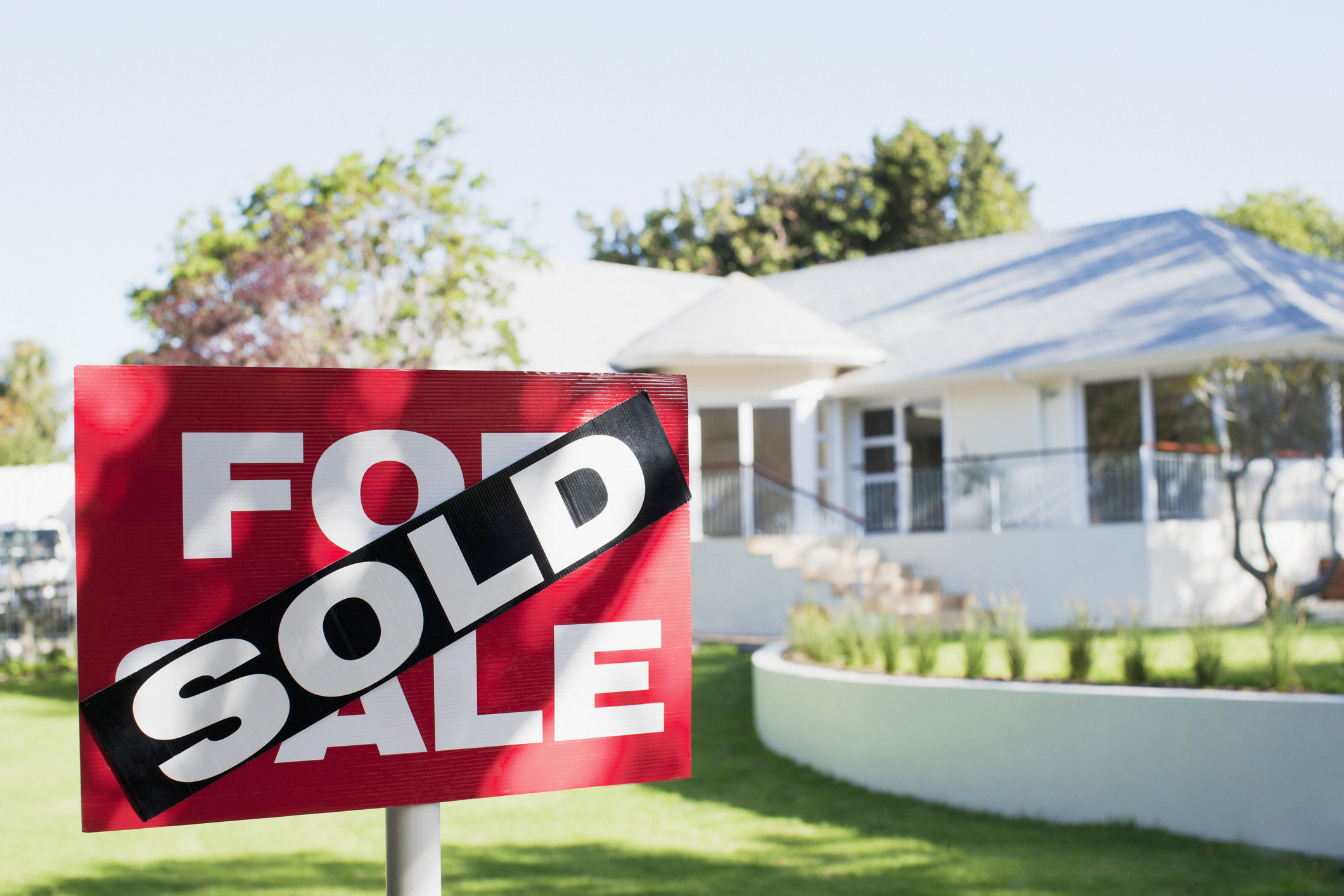 Close-up of a "Sold" sign in front of a house with a well-maintained lawn and trees