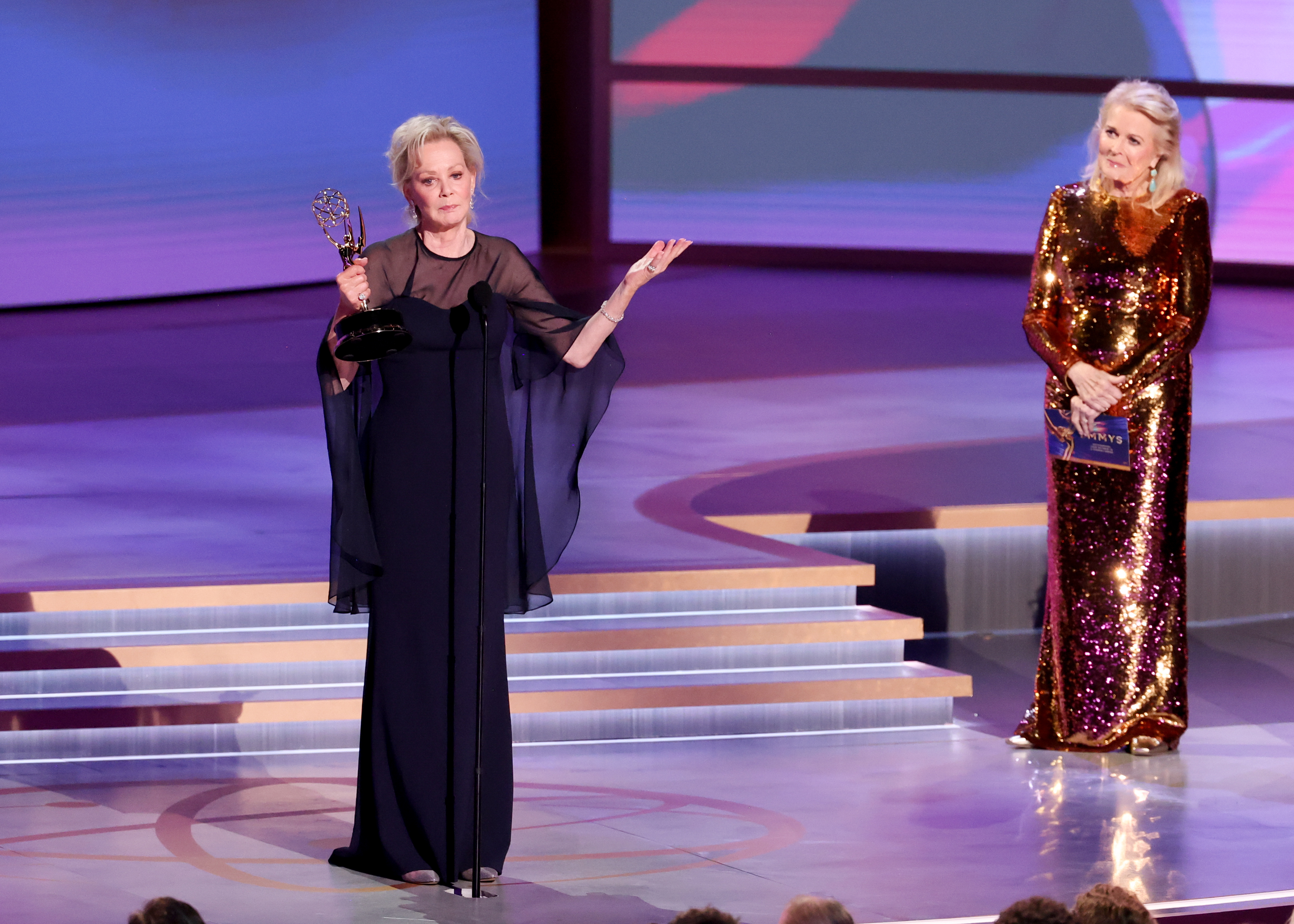 Jean Smart accepts an award on stage, wearing a sleek gown, while Candice Bergen stands nearby in a dazzling sequin dress holding an envelope
