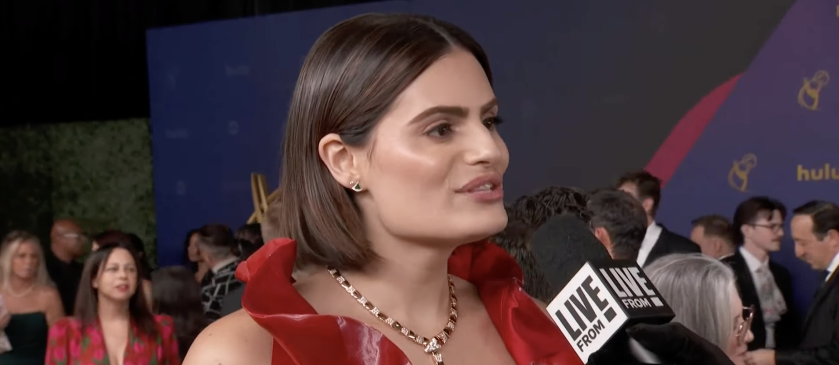 Nava Mau on the red carpet, speaking into a "Live From E!" microphone. She is wearing a sleeveless dress with a structured neckline and a beaded necklace