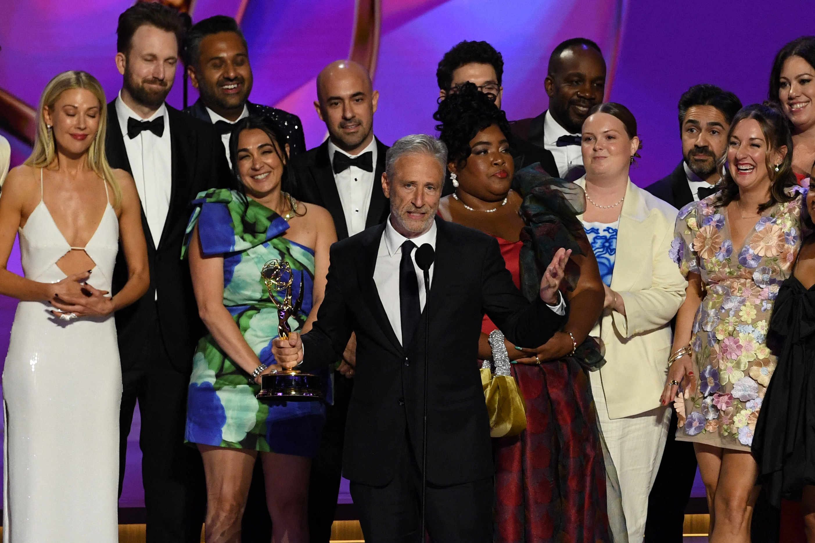 Jon Stewart, holding an Emmy, stands on stage with the "The Daily Show with Trevor Noah" cast and crew, wearing formal attire at an award ceremony