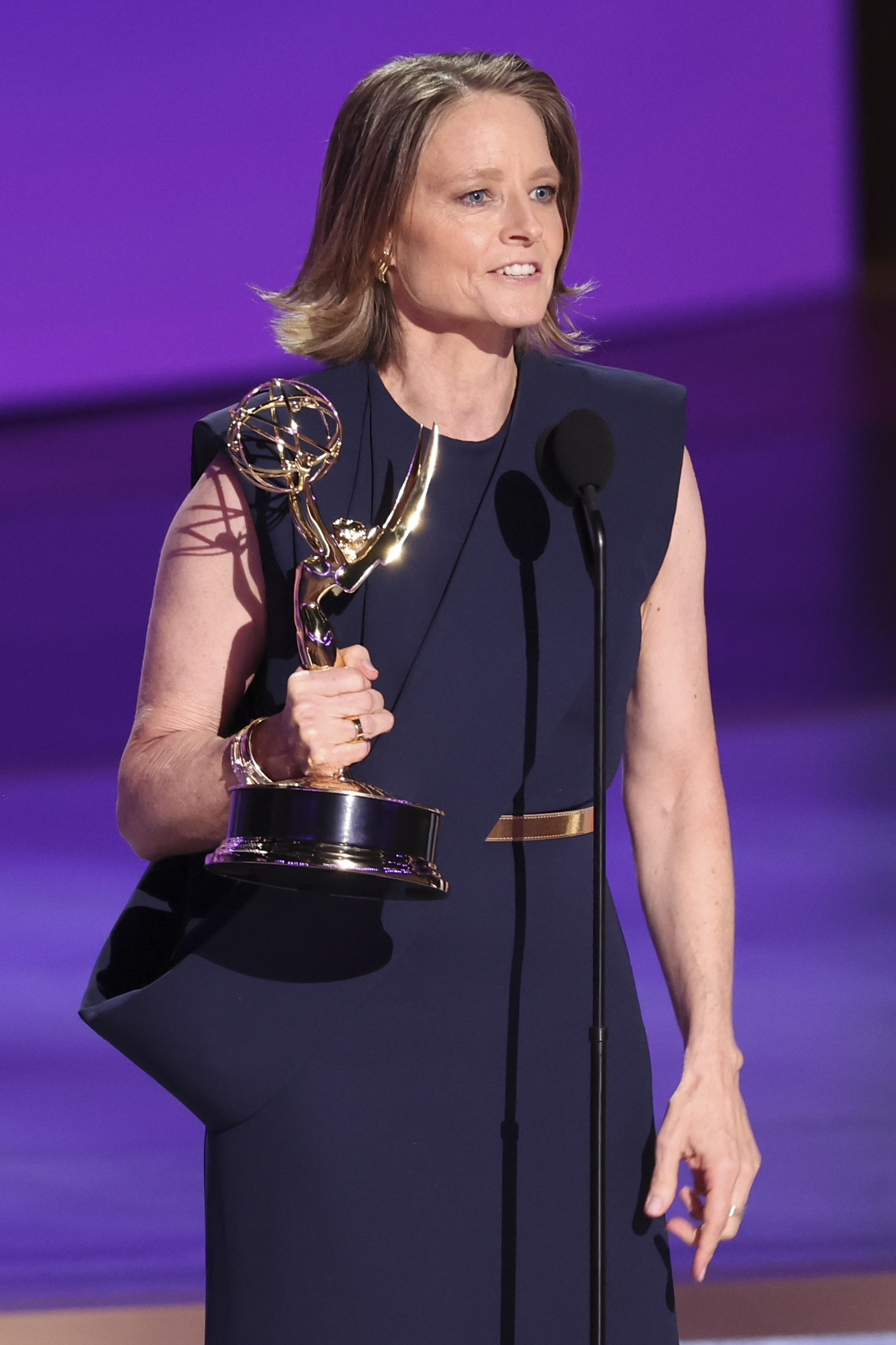 Jodie Foster stands on stage in elegant attire, holding an Emmy Award and speaking into a microphone