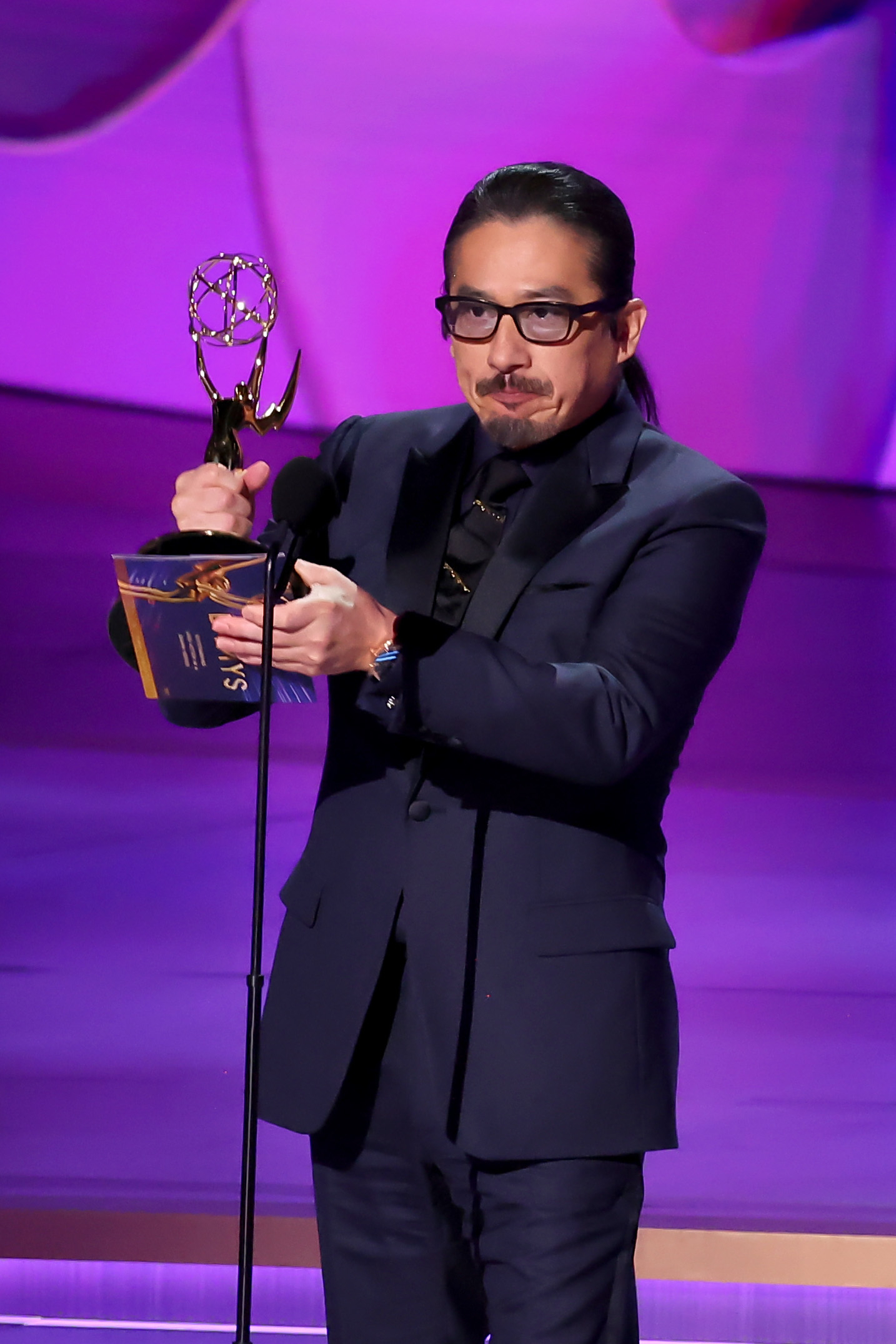 Hiroyuki Sanada holds an Emmy award and speaks into a microphone on stage, wearing a dark suit and glasses