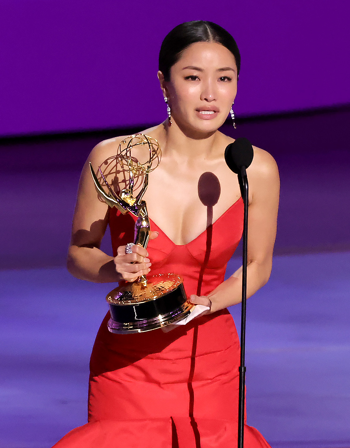 Anna Sawai, in an elegant gown, holds an Emmy Award while speaking at a microphone on stage