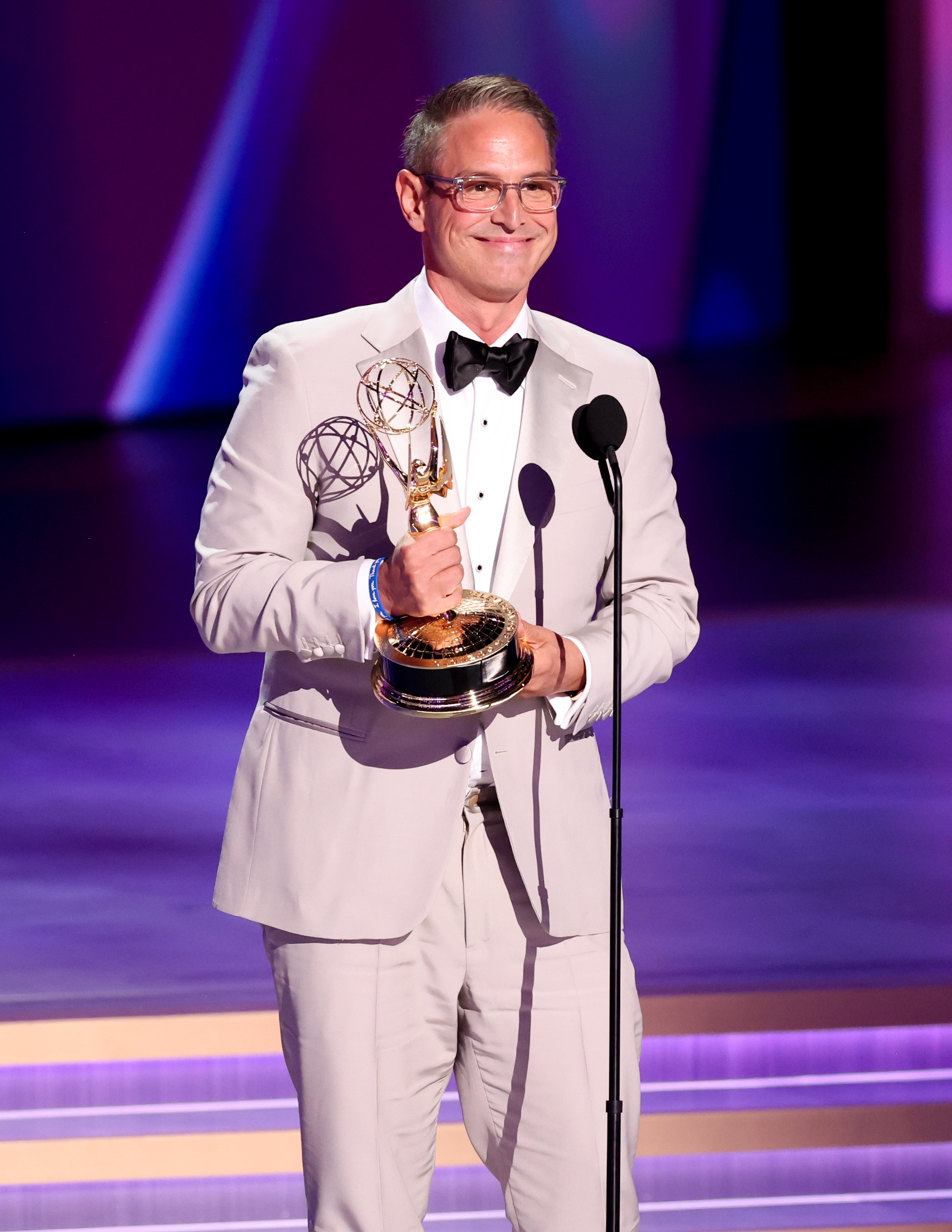 Man in a light suit and bow tie holds an Emmy award, standing on stage at an awards ceremony
