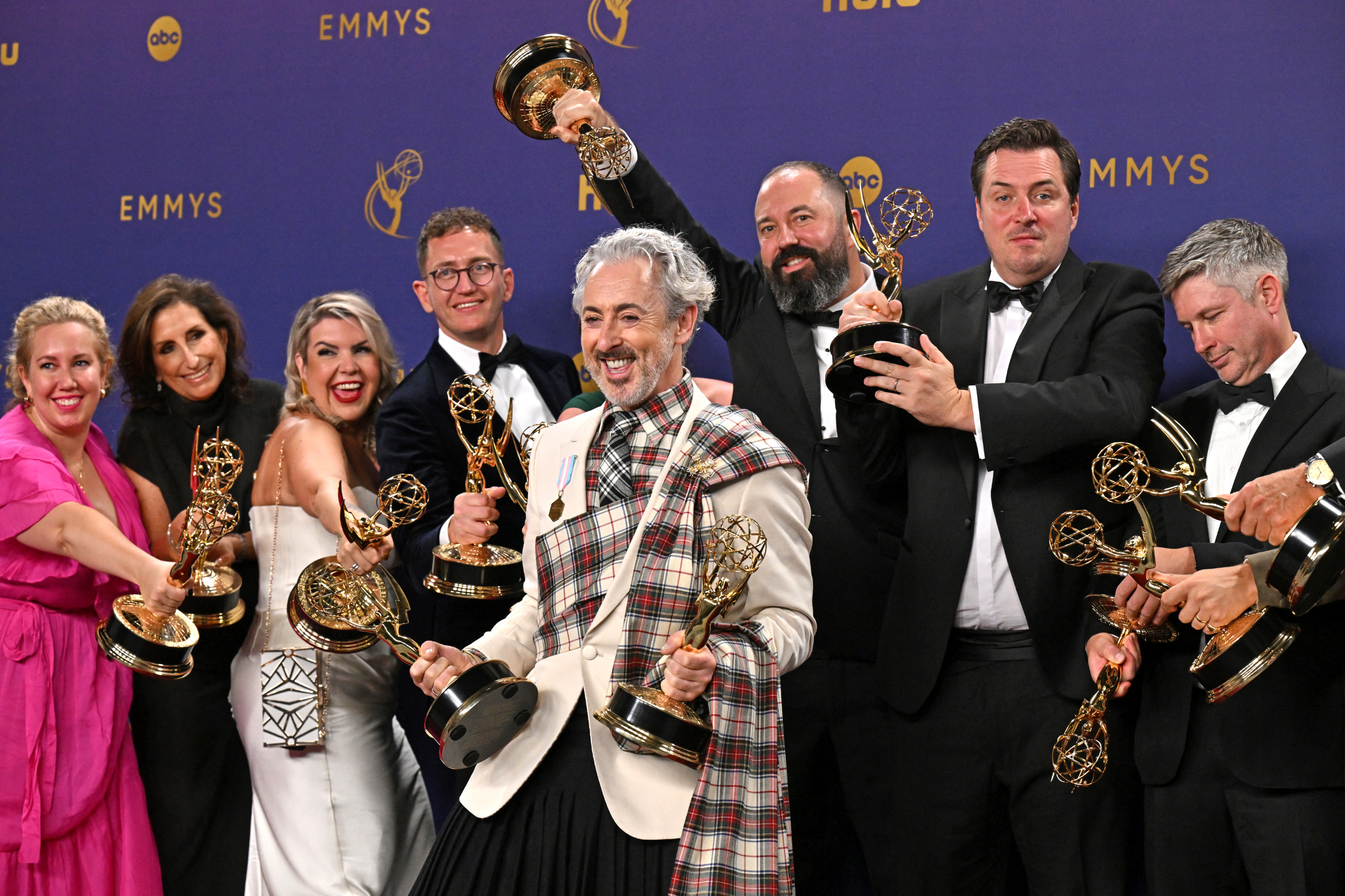 A cast at an Emmy awards event celebrates on stage with their awards. They are dressed in formal attire and smiling proudly