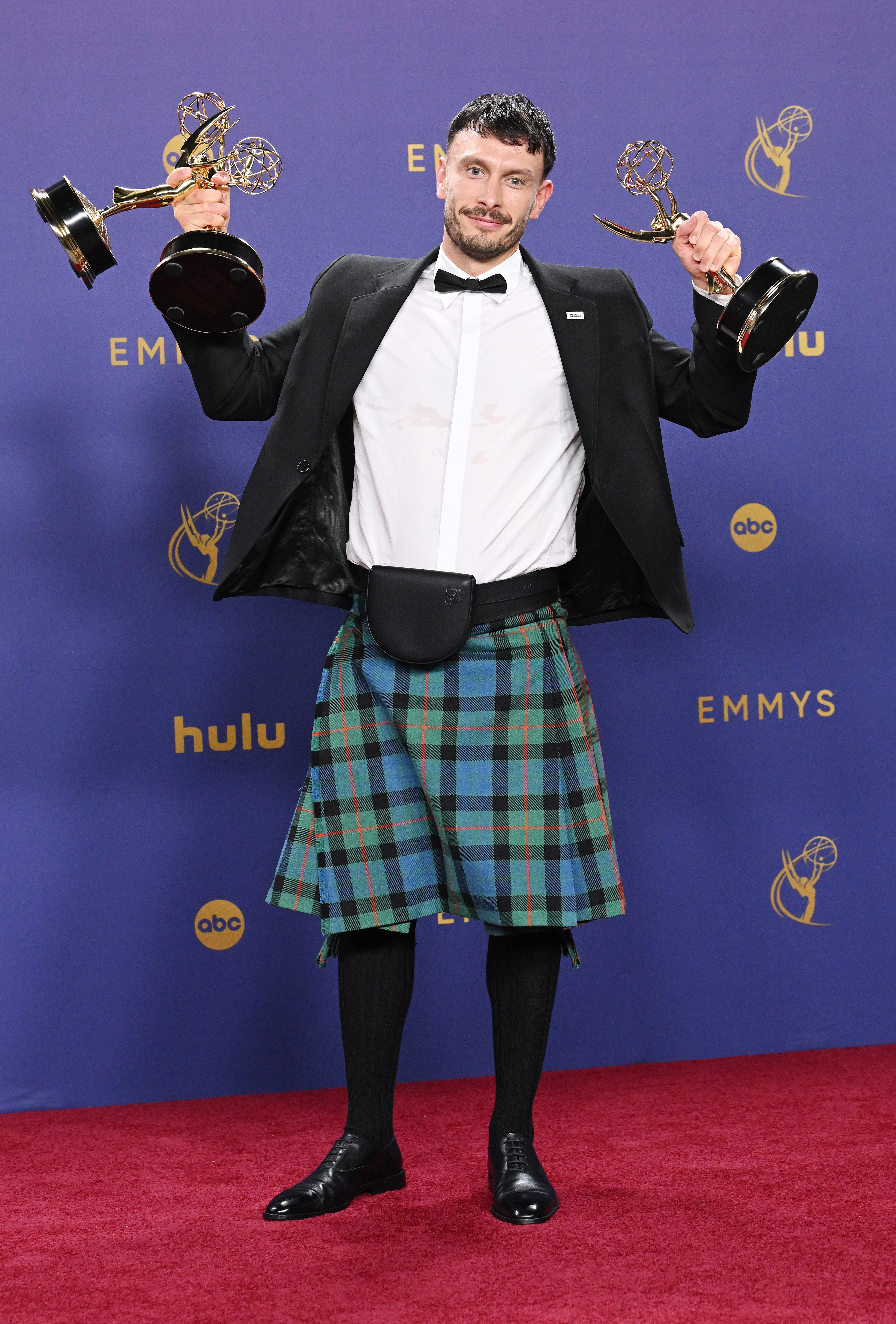 Actor Richard Gadd poses on the Emmys red carpet holding two awards, wearing a tuxedo jacket, white shirt, bow tie, and a tartan kilt