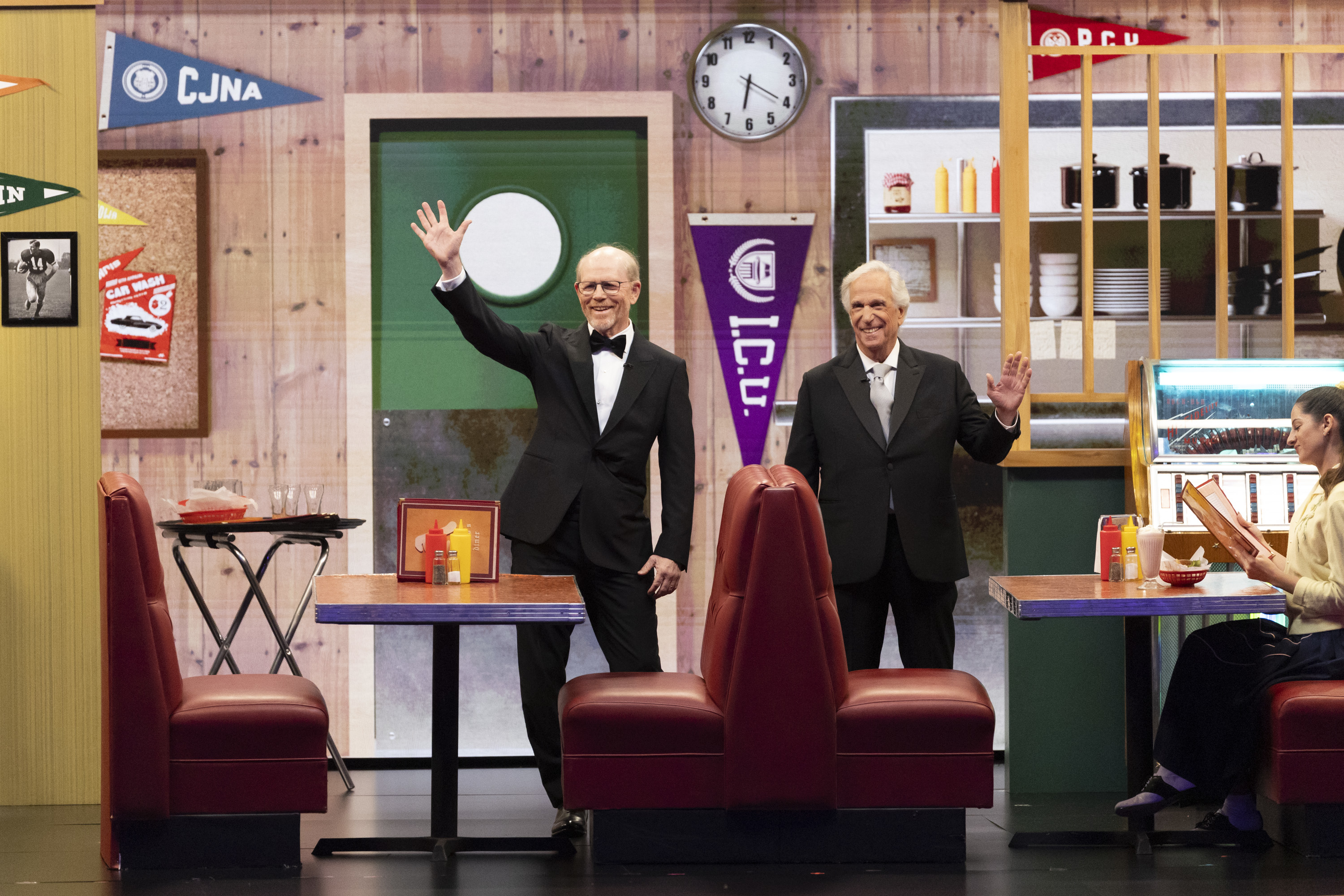 Christopher Lloyd and Michael J. Fox, both in suits, wave from a TV set resembling a classic diner, promoting a reunion event
