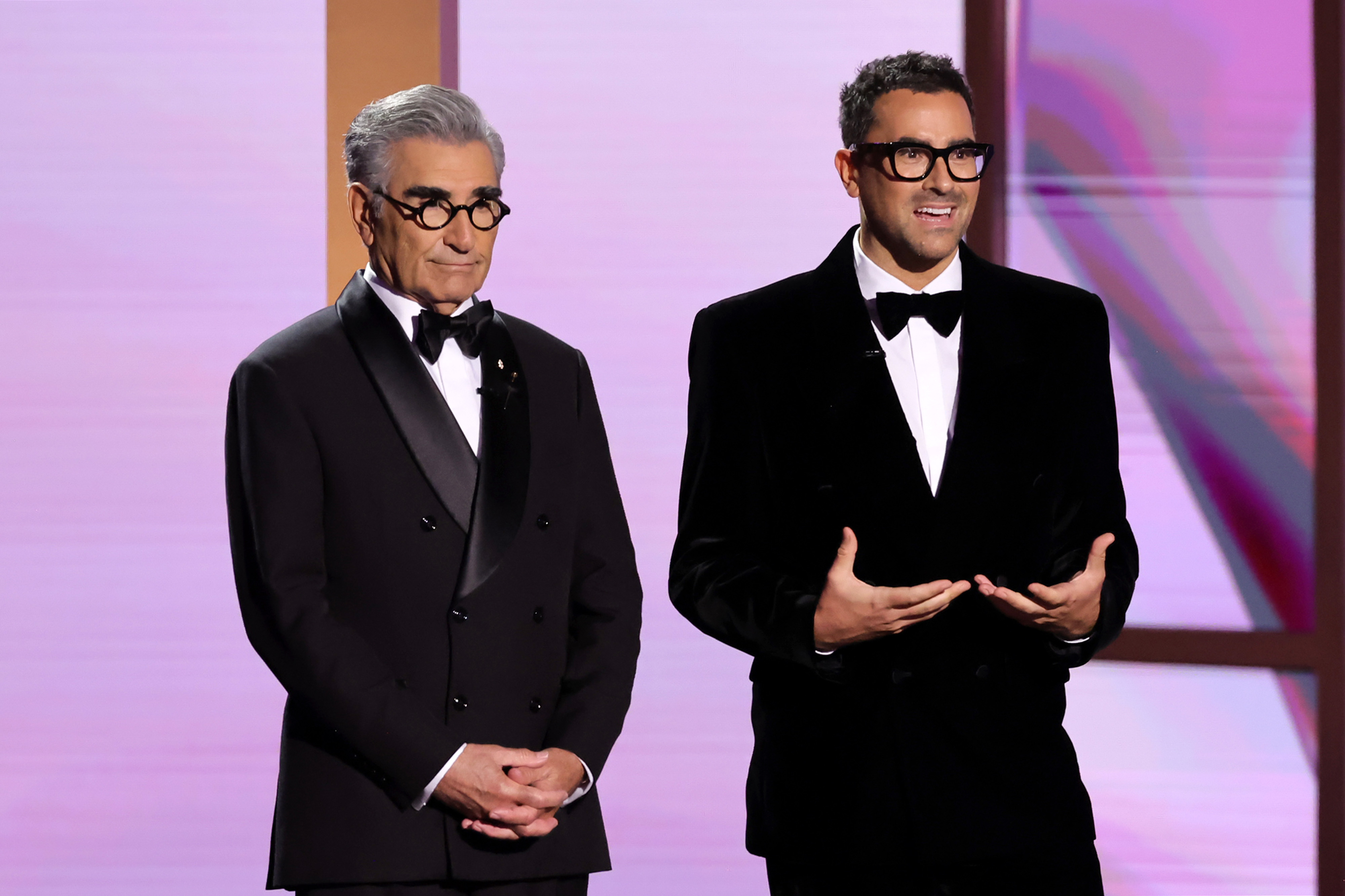 Eugene Levy and Dan Levy in tuxedos, standing on stage, addressing the audience at an event