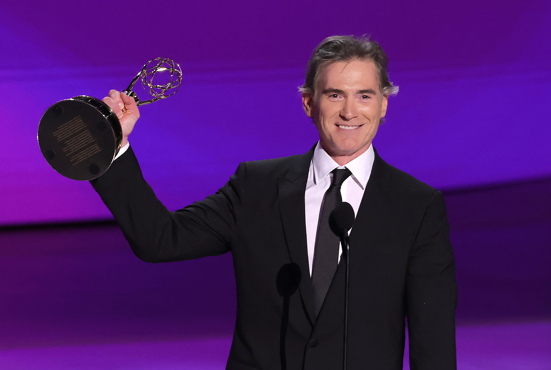 Billy Crudup on stage, smiling and holding up an Emmy award while wearing a suit and tie