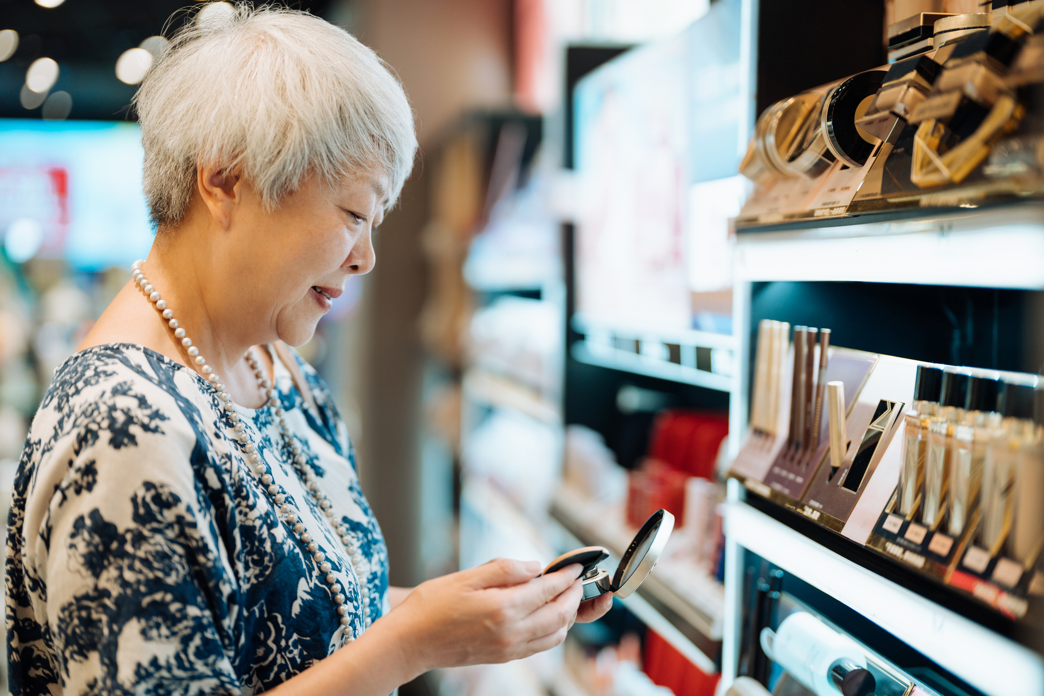 An elderly woman with short white hair, wearing a floral blouse and pearl necklace, smiles while looking at a compact in a makeup store