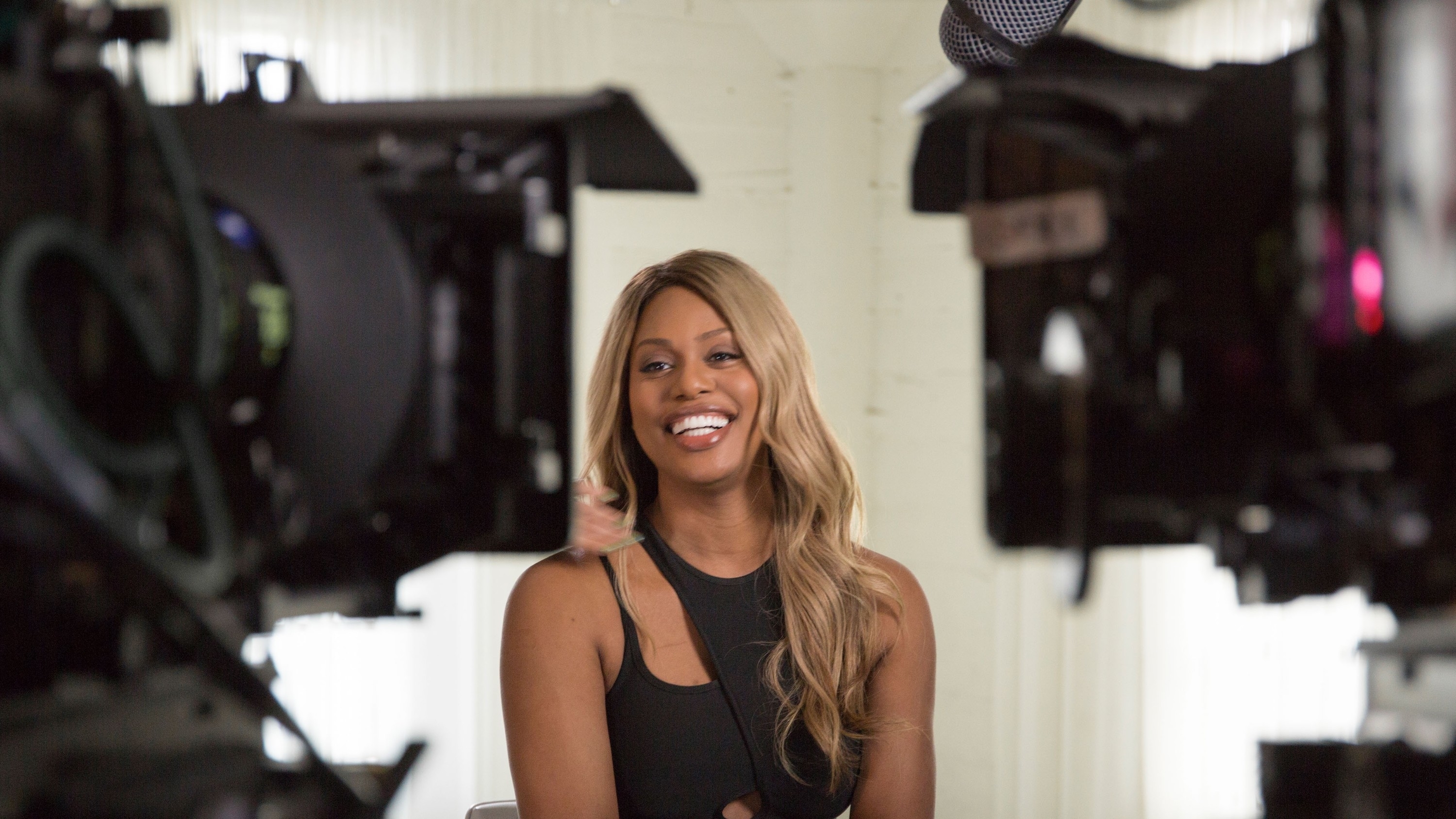 Laverne Cox sits smiling between two video cameras during an interview. She is wearing a black sleeveless top
