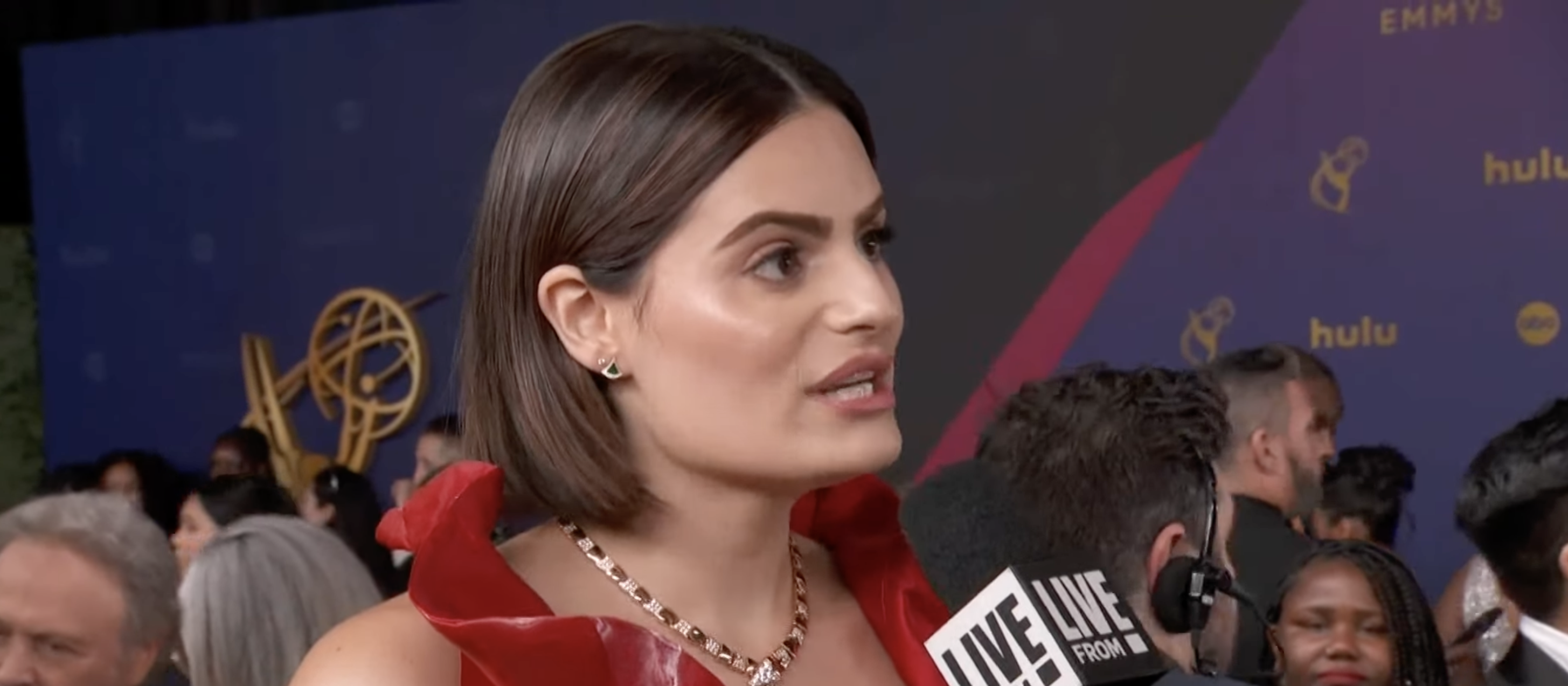 Nava speaking into a microphone on the red carpet at the Emmys. She has short hair and wears a glamorous, ruffled dress. People and media are in the background