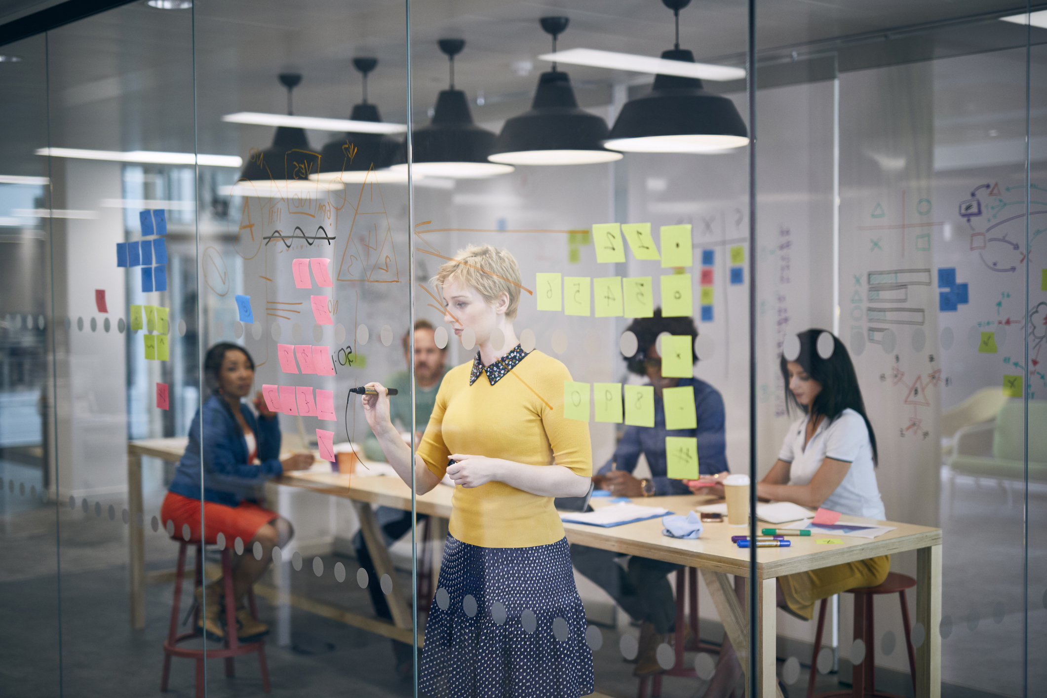 A woman writes on a glass board with sticky notes, while three colleagues in casual attire brainstorm at a table in the background