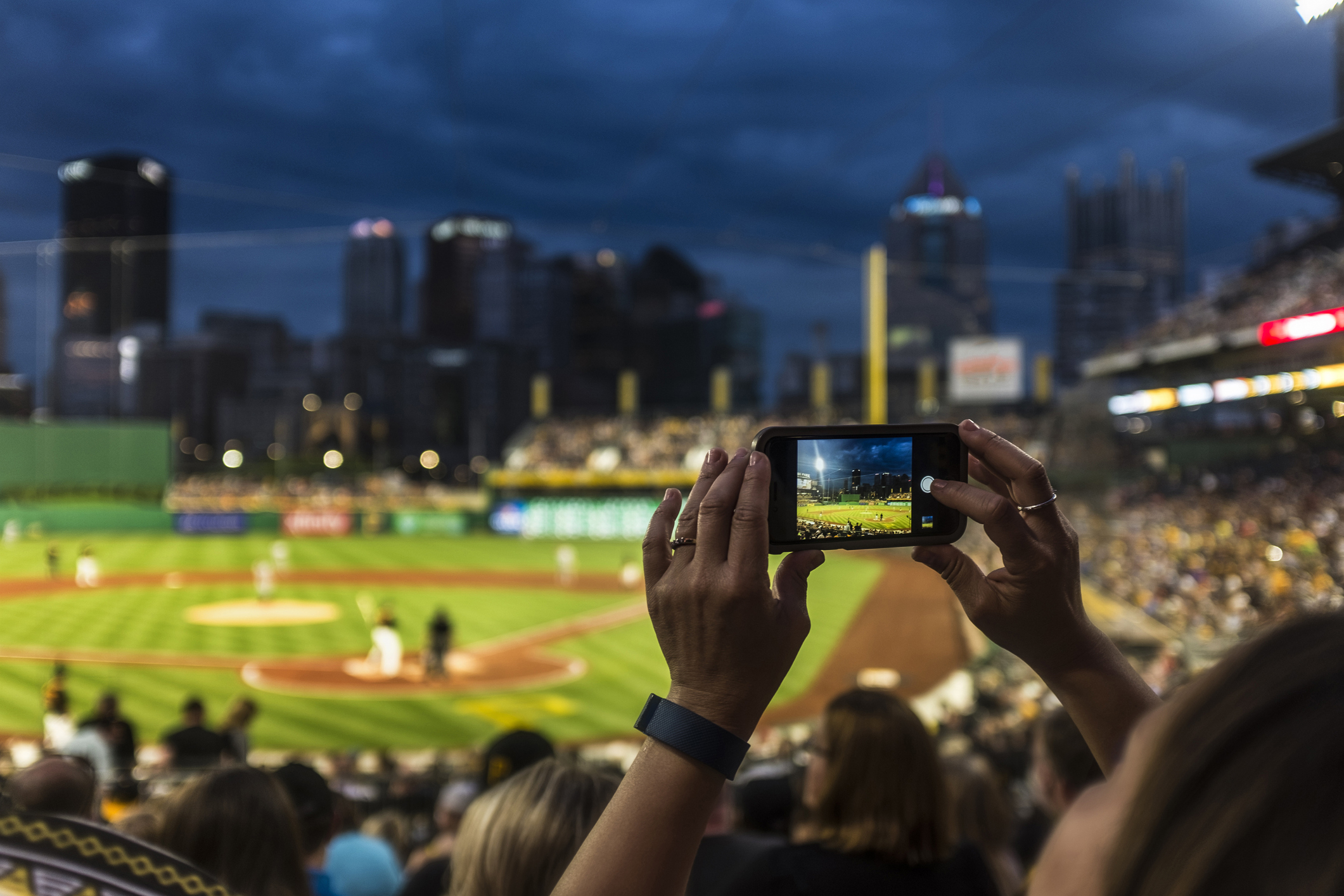 A person holds up a phone to take a photo of a baseball game in a packed stadium at dusk