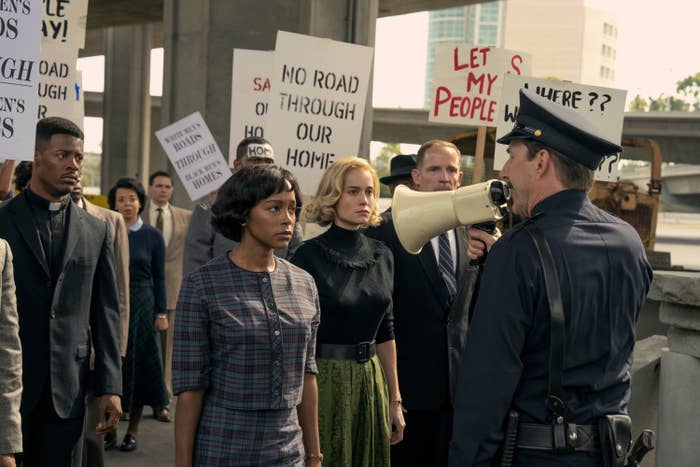 Actors, including Naomi,  in a protest scene from "Just Mercy," holding signs like "No road through our homes" while a policeman addresses them with a megaphone