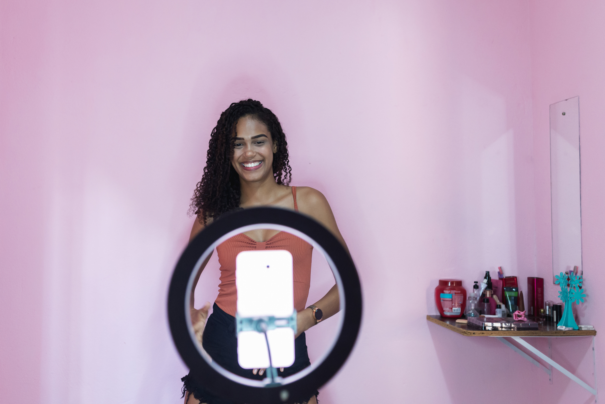 Woman posing in front of a ring light and smartphone, smiling, with beauty products on a shelf behind her