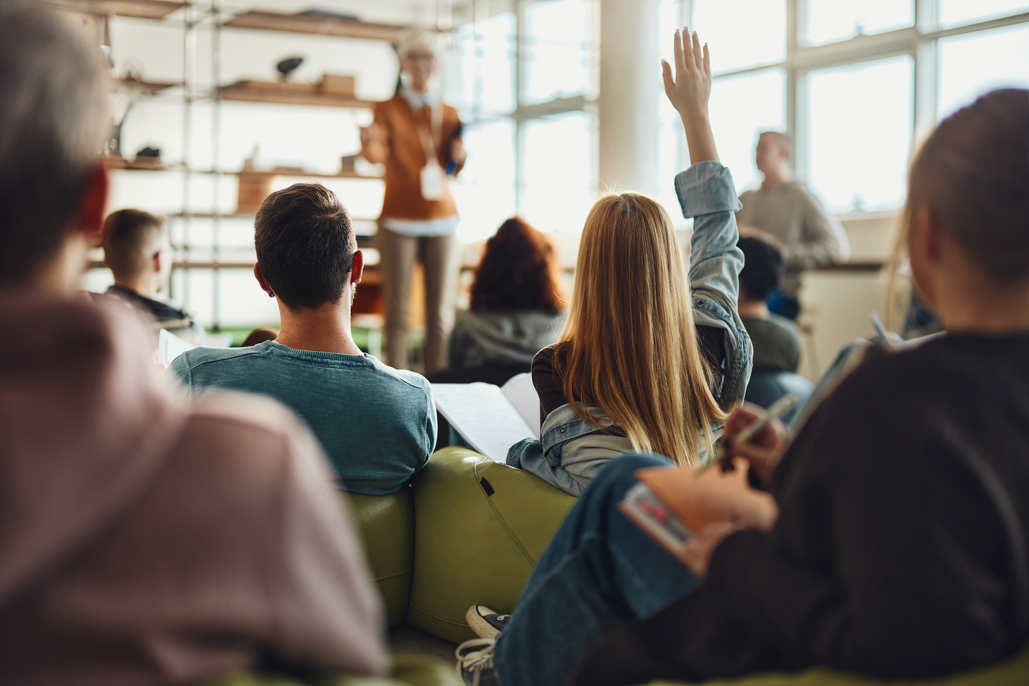 Students in a classroom, one student raising their hand to ask a question, facing an instructor at the front of the room