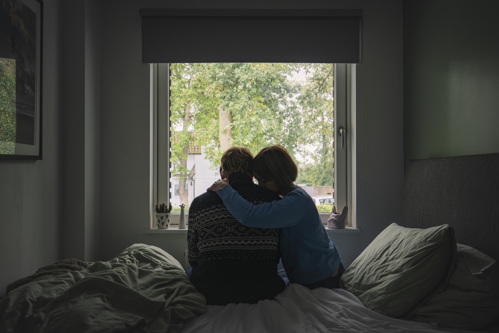 Parents sit closely together on a bed, looking out of a window at a green, leafy scene outside. One has an arm wrapped around the other in a comforting gesture