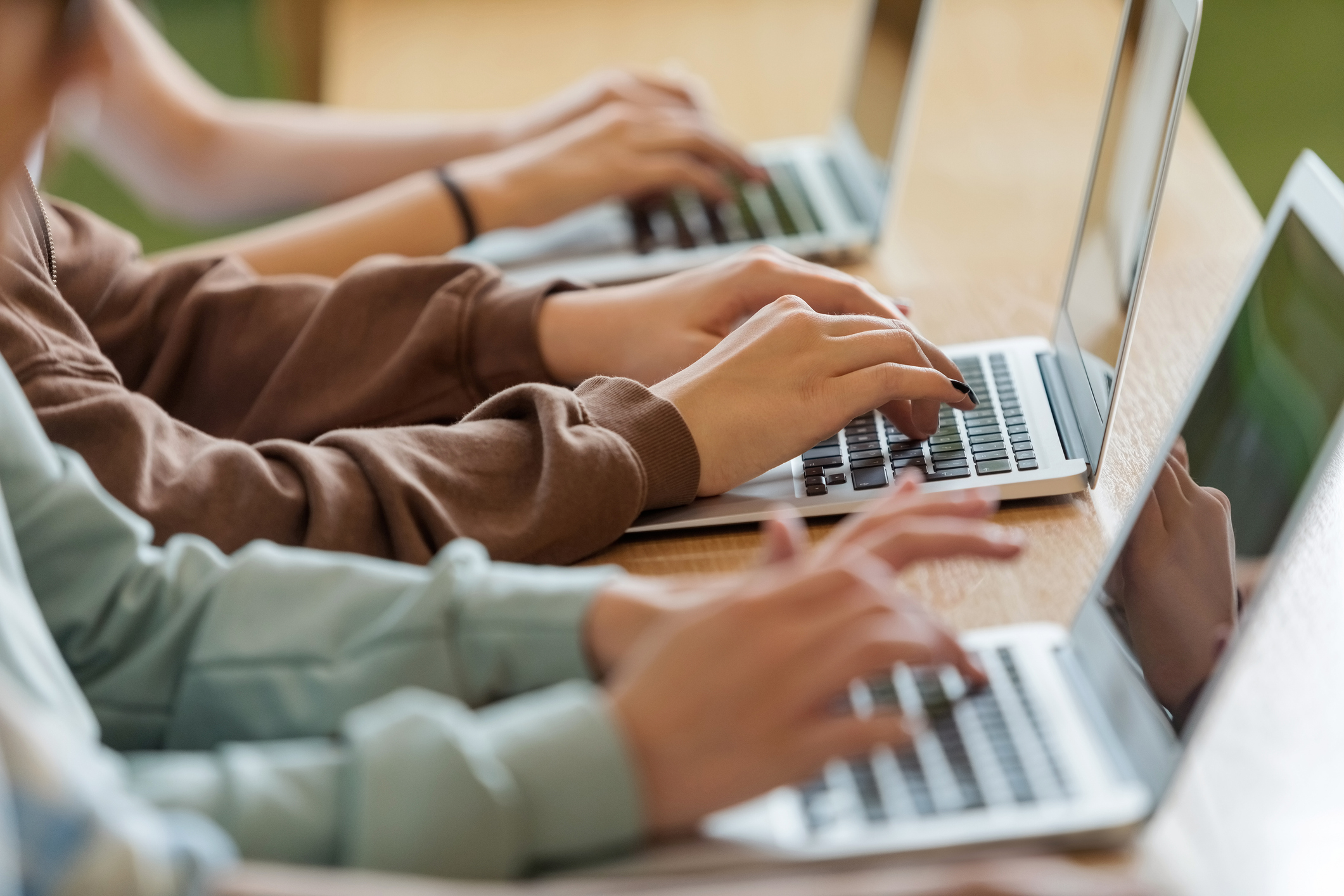 Multiple people type on laptops, seated side by side at a table. Only their hands and forearms are visible in the image