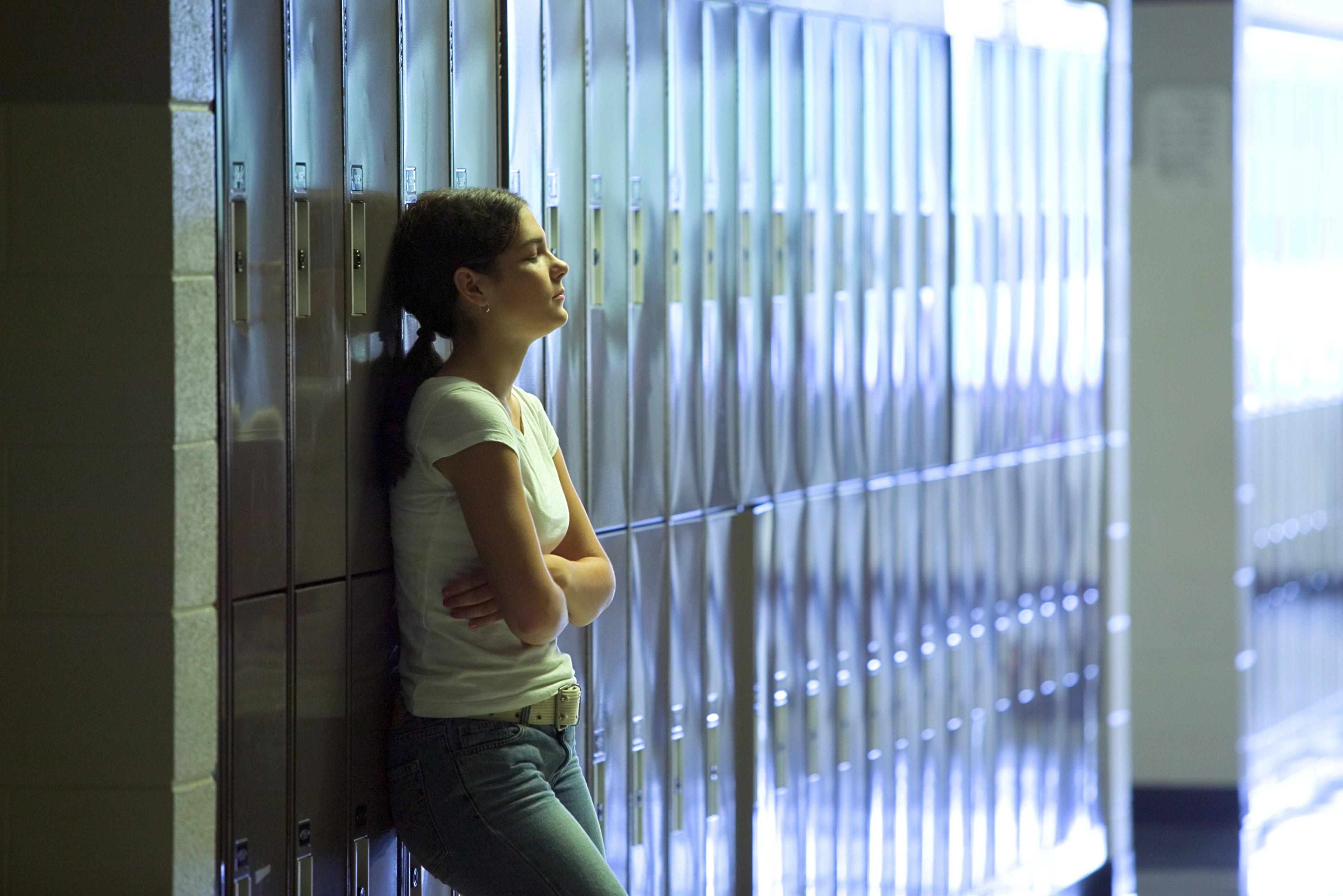 A person leans against school lockers, arms folded, eyes closed, in a seemingly pensive or contemplative state. Light shines through a distant window